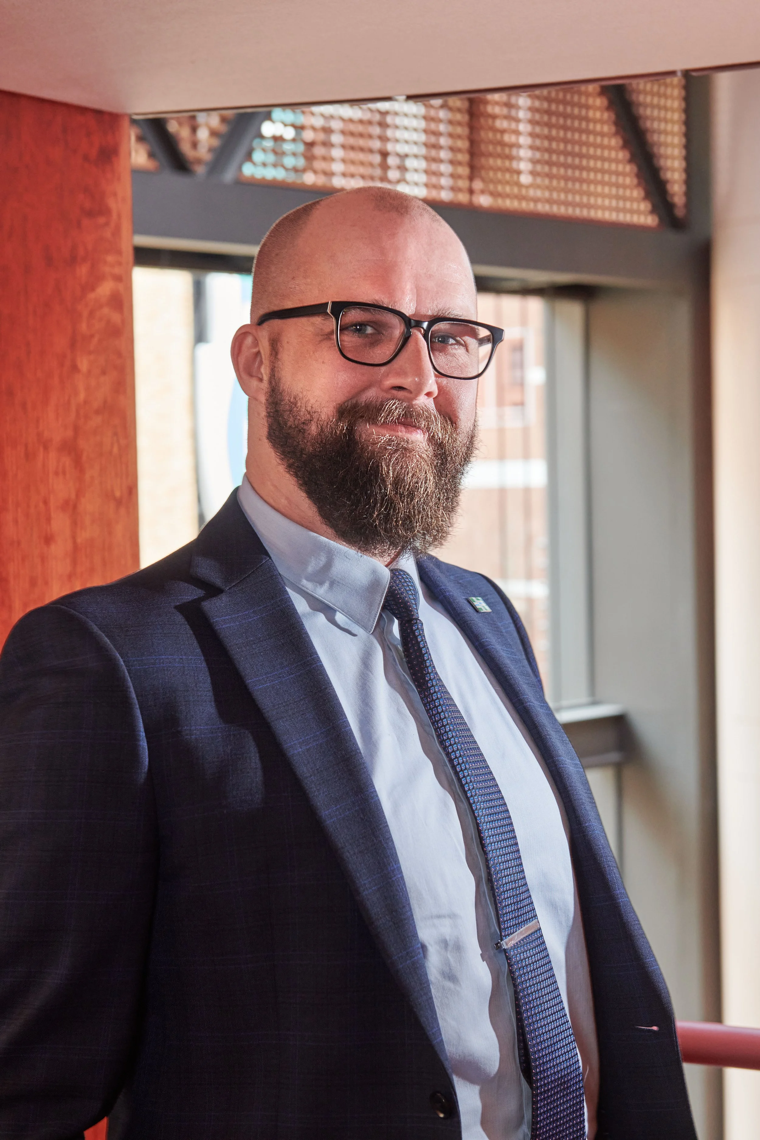 A man with a beard and glasses dressed in a suit, standing indoors near a window with sunlight, posing confidently.