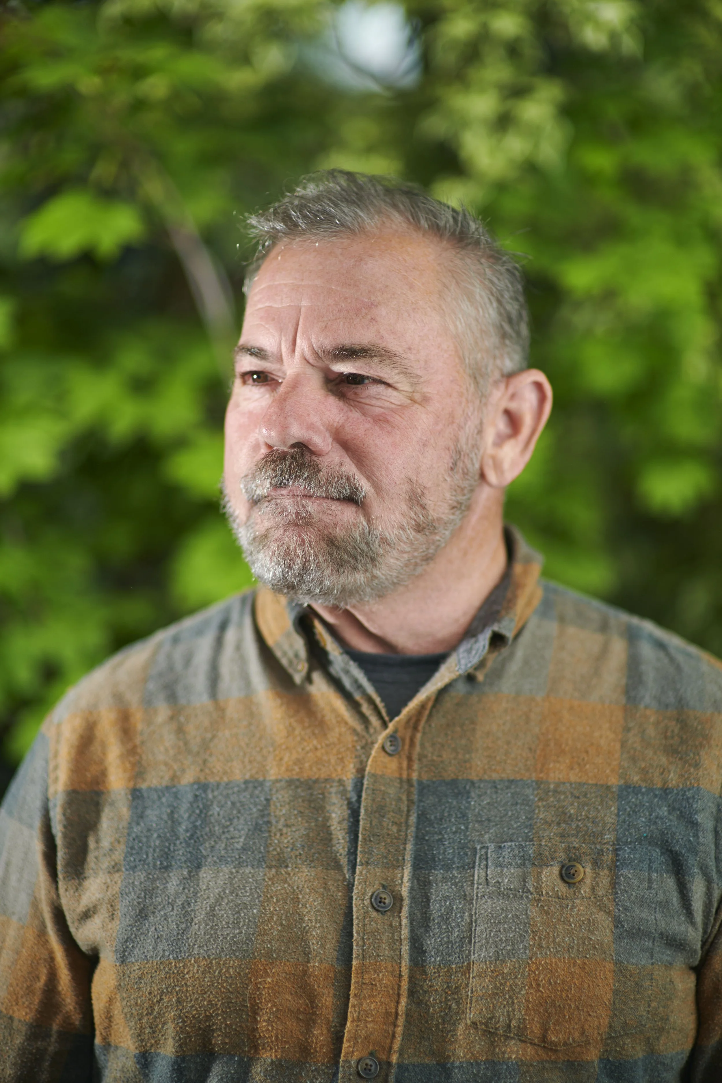 A middle-aged man with gray hair and a beard, wearing a plaid shirt, stands outdoors with green foliage in the background.