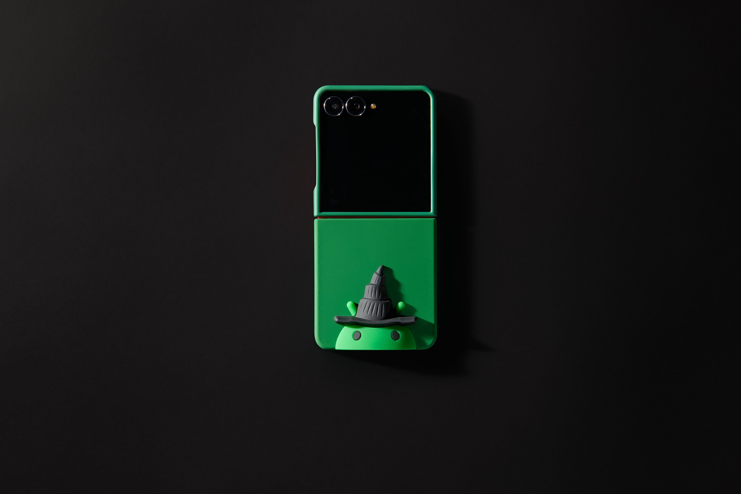 A green Android phone case with Bot wearing a Wicked witch hat on the phone case against a black background. The case is merch for the movie Wicked.