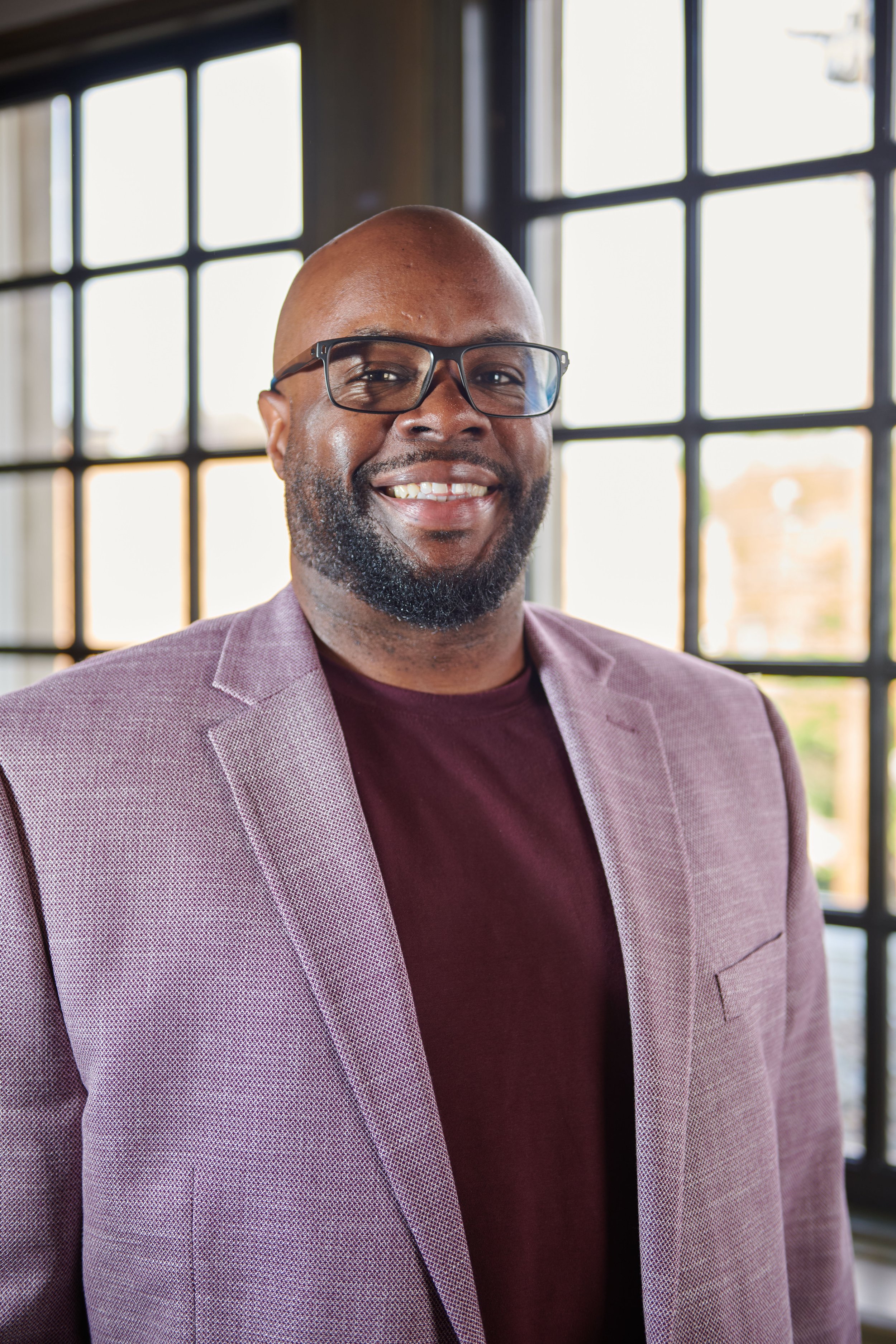 A portrait of a smiling man with glasses, beard, and shaved head, wearing a purple blazer and maroon shirt, standing indoors in front of large windows.