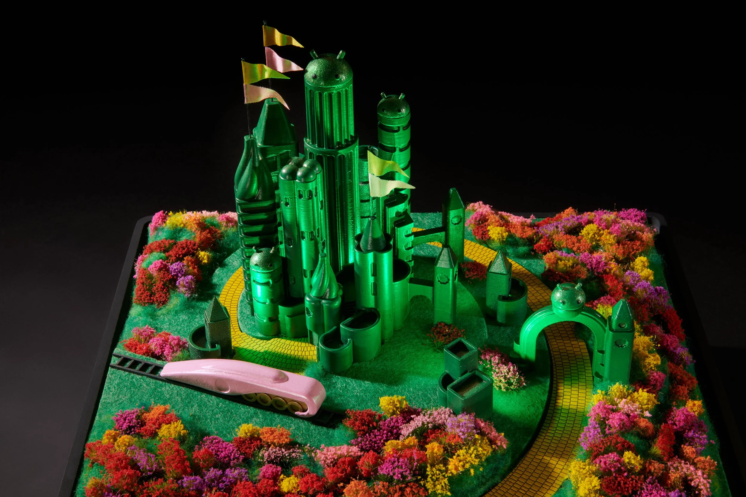 Colorful 3D model of Oz from the movie Wicked for Good with green skyscrapers, pink and yellow flags, and flower-filled landscape on a black background.