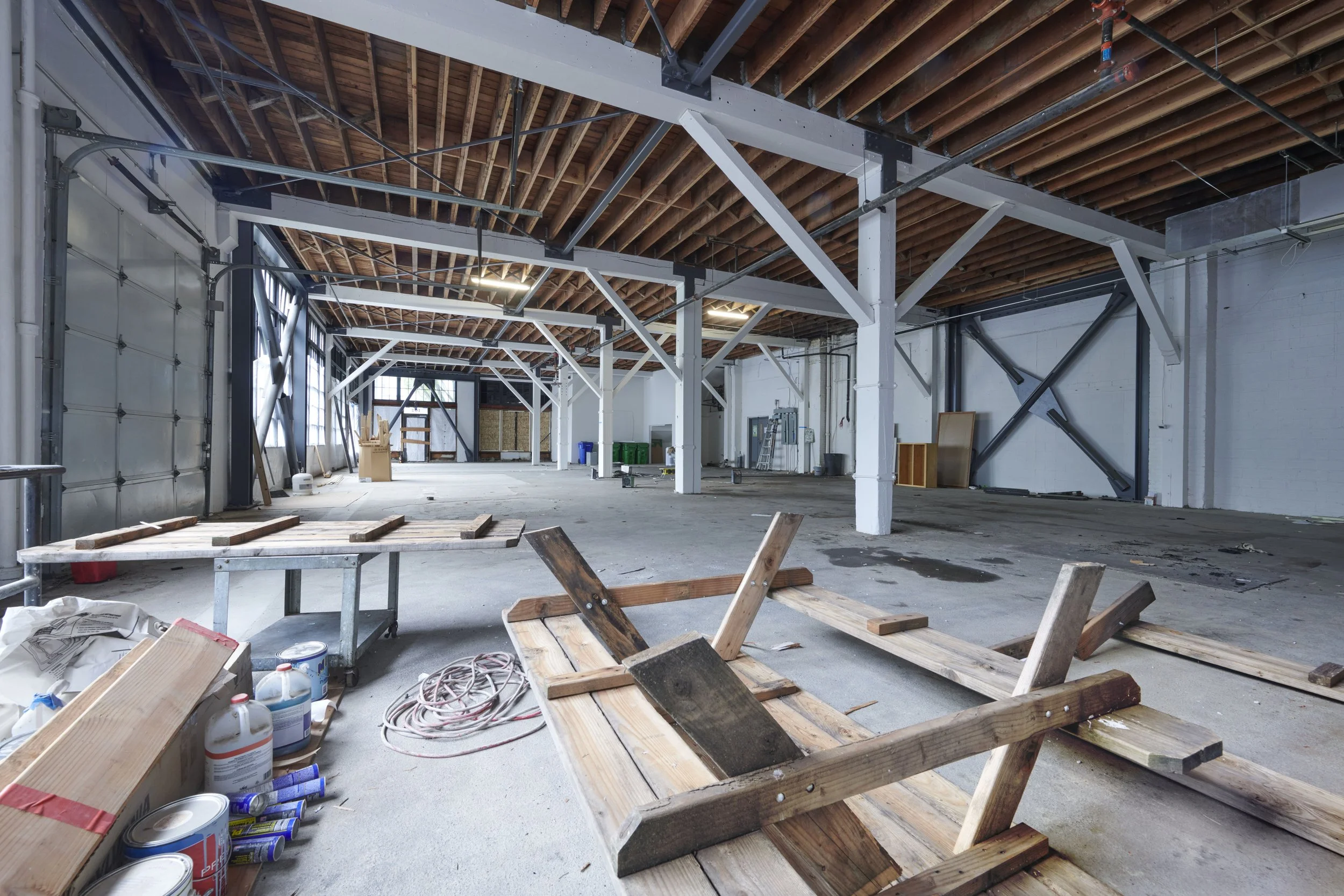 Interior of a warehouse or industrial building under construction with exposed wooden ceiling beams, metal support columns, and construction materials scattered on the floor.