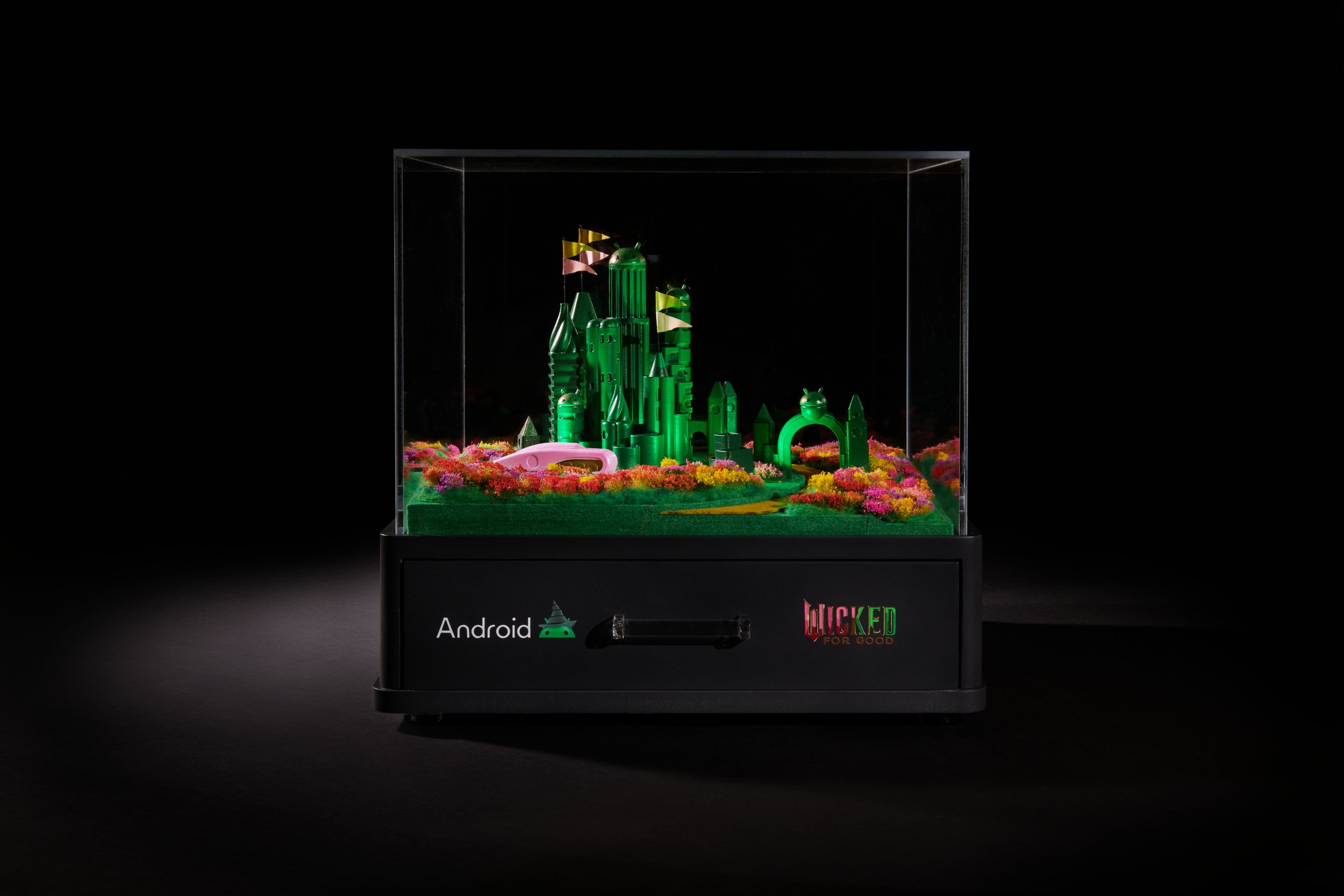 Miniature green cityscape with buildings, trees, and flags in a glass display case, labeled 'Android' and 'Wicked for Good' on the front.