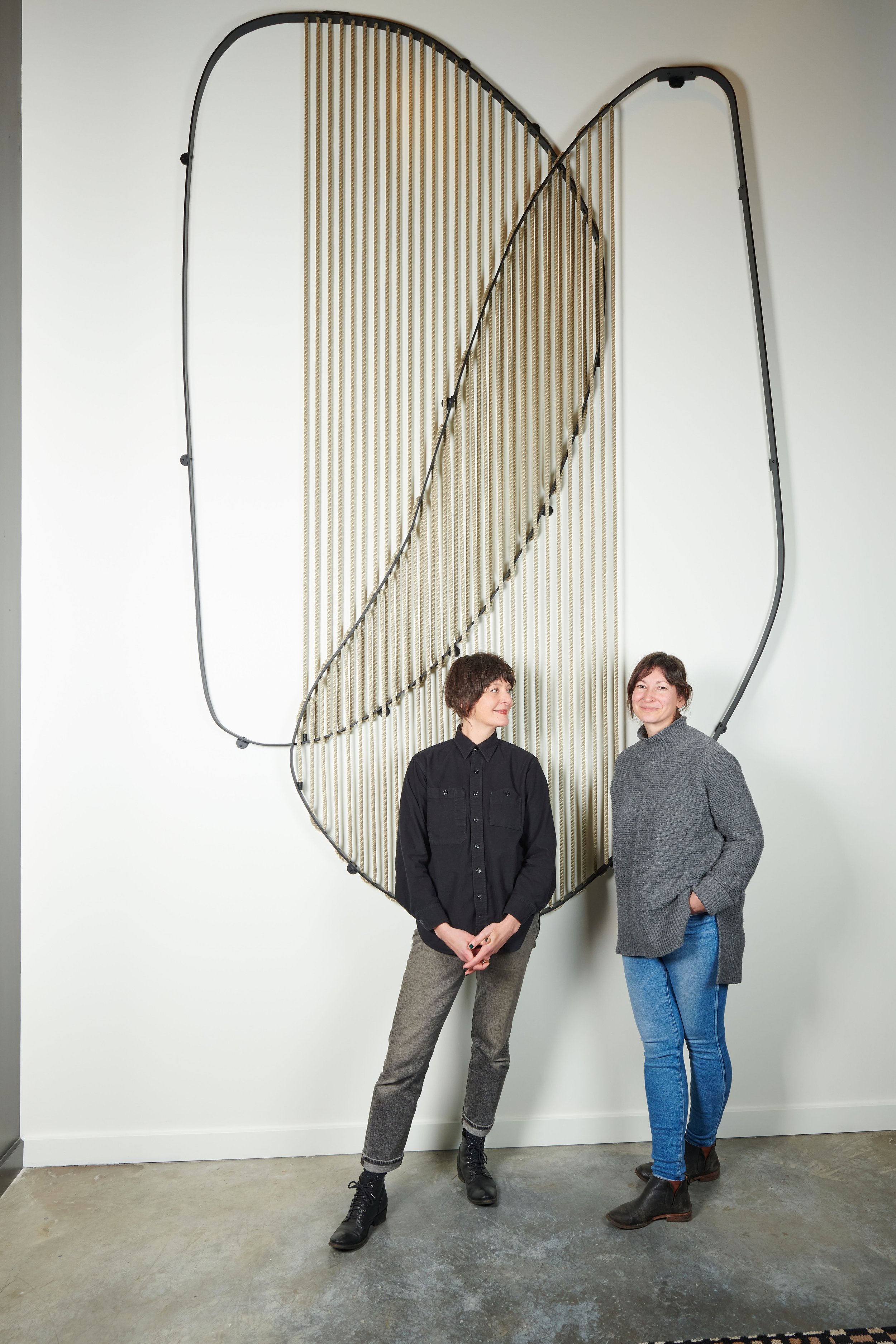 Two women standing in front of a wall art installation with a large leaf outline made of black metal and vertical gold-colored rods.