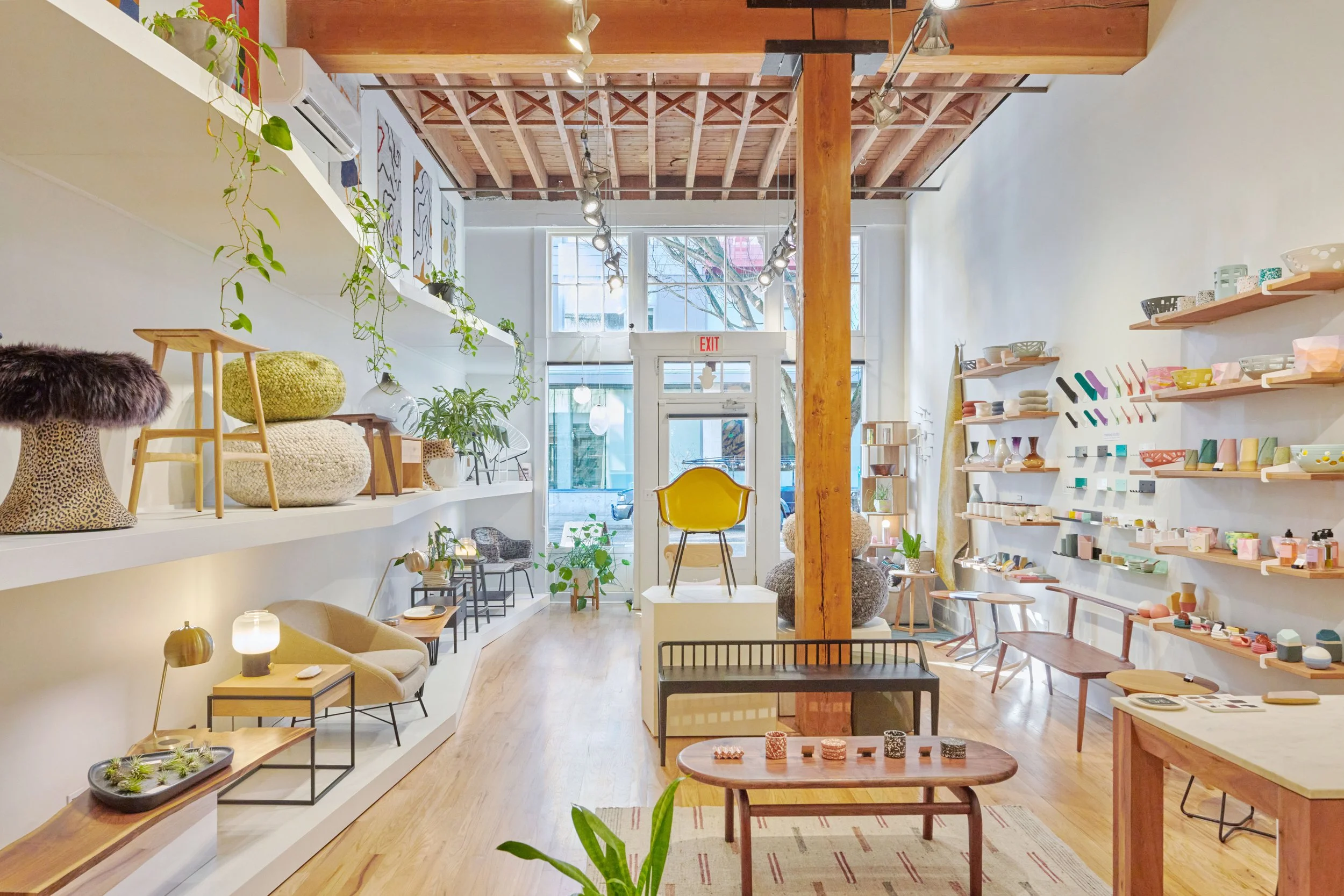 Interior of a modern retail store with display shelves holding ceramics, decor items, and plants. Items are arranged on white shelves on the walls and on tables. Large windows at the back let in natural light, and the ceiling features exposed wooden 
