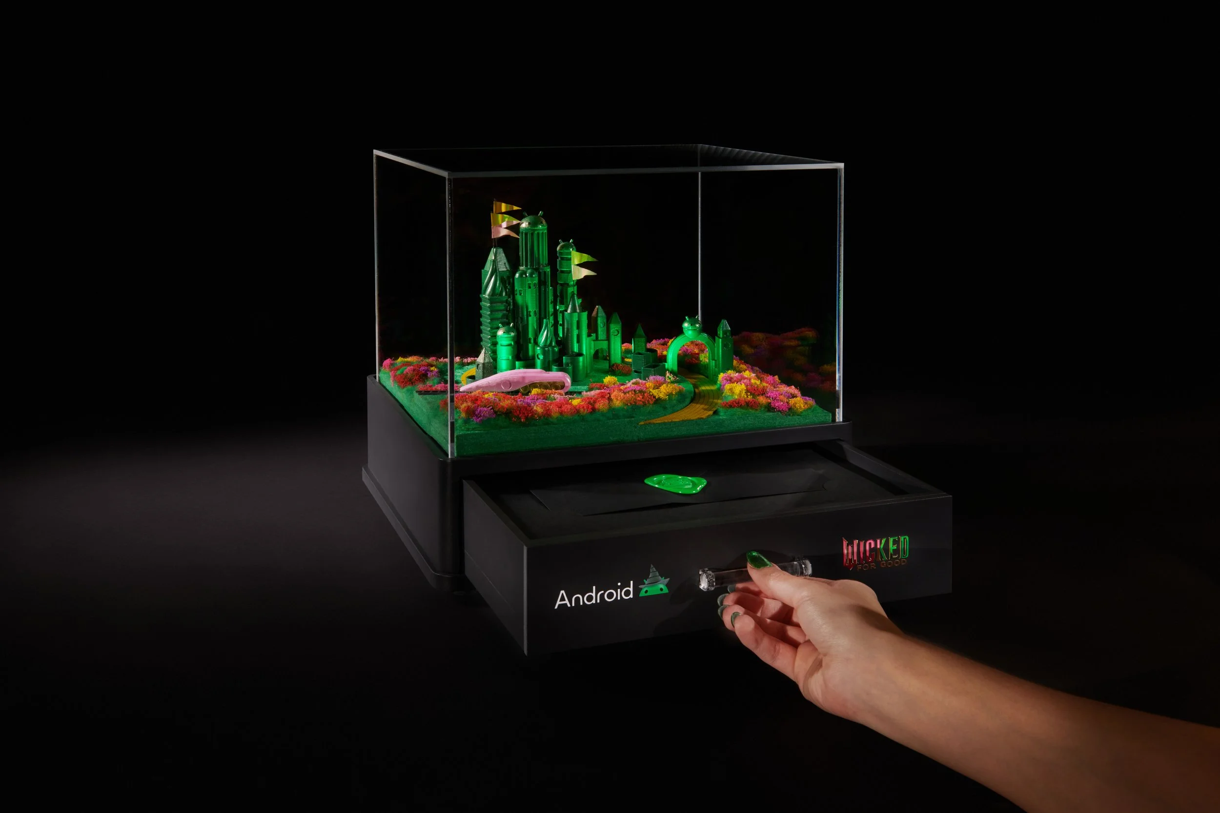 A person's hand with painted nails turning a knob on a black box labeled Android, revealing a miniature green castle with flags inside a glass display case. The castle is surrounded by colorful flowers, and the background is black.