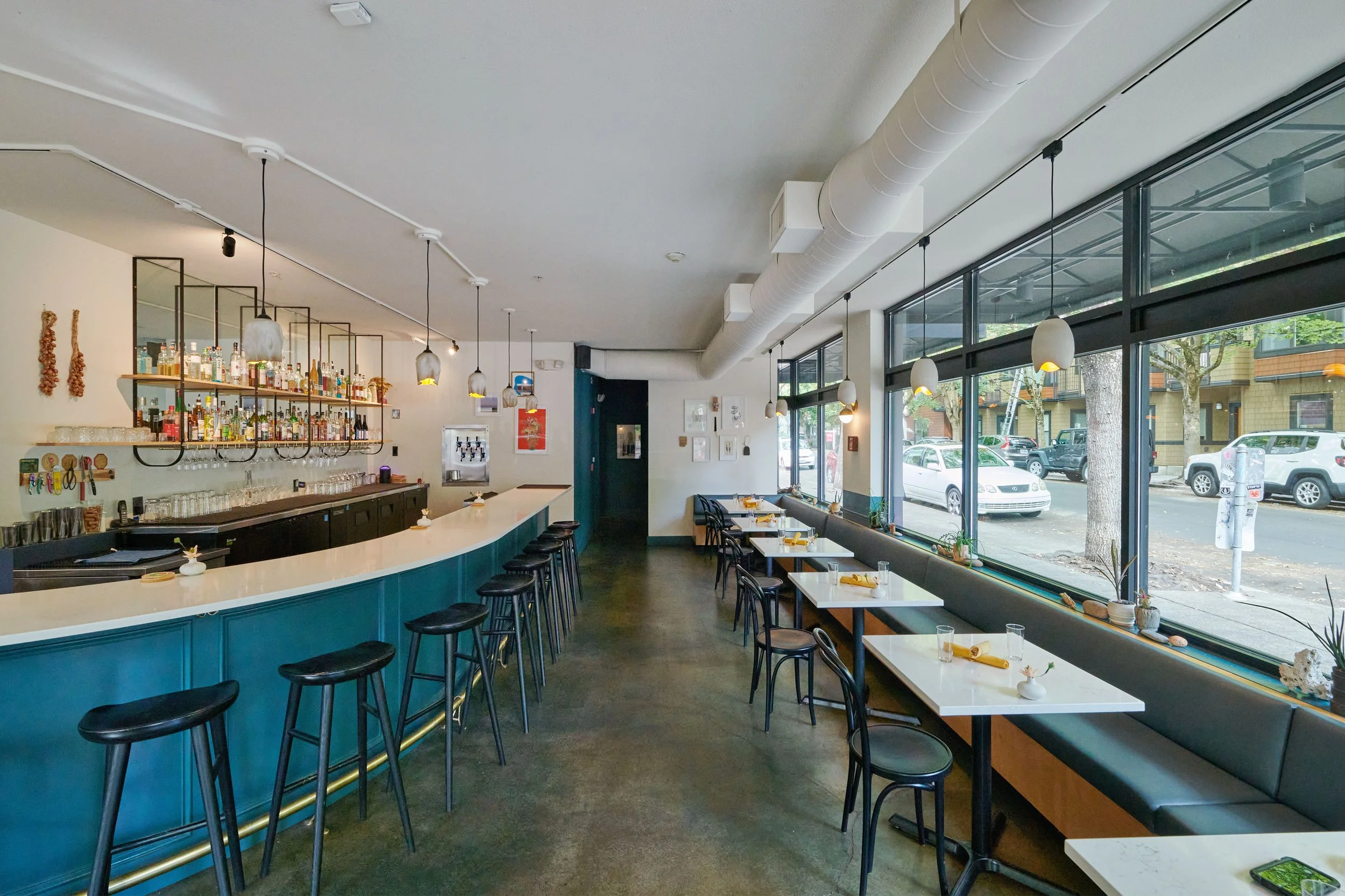 Modern restaurant interior with white tables, black chairs, large windows, pendant lighting, and a bar with alcohol bottles on shelves.