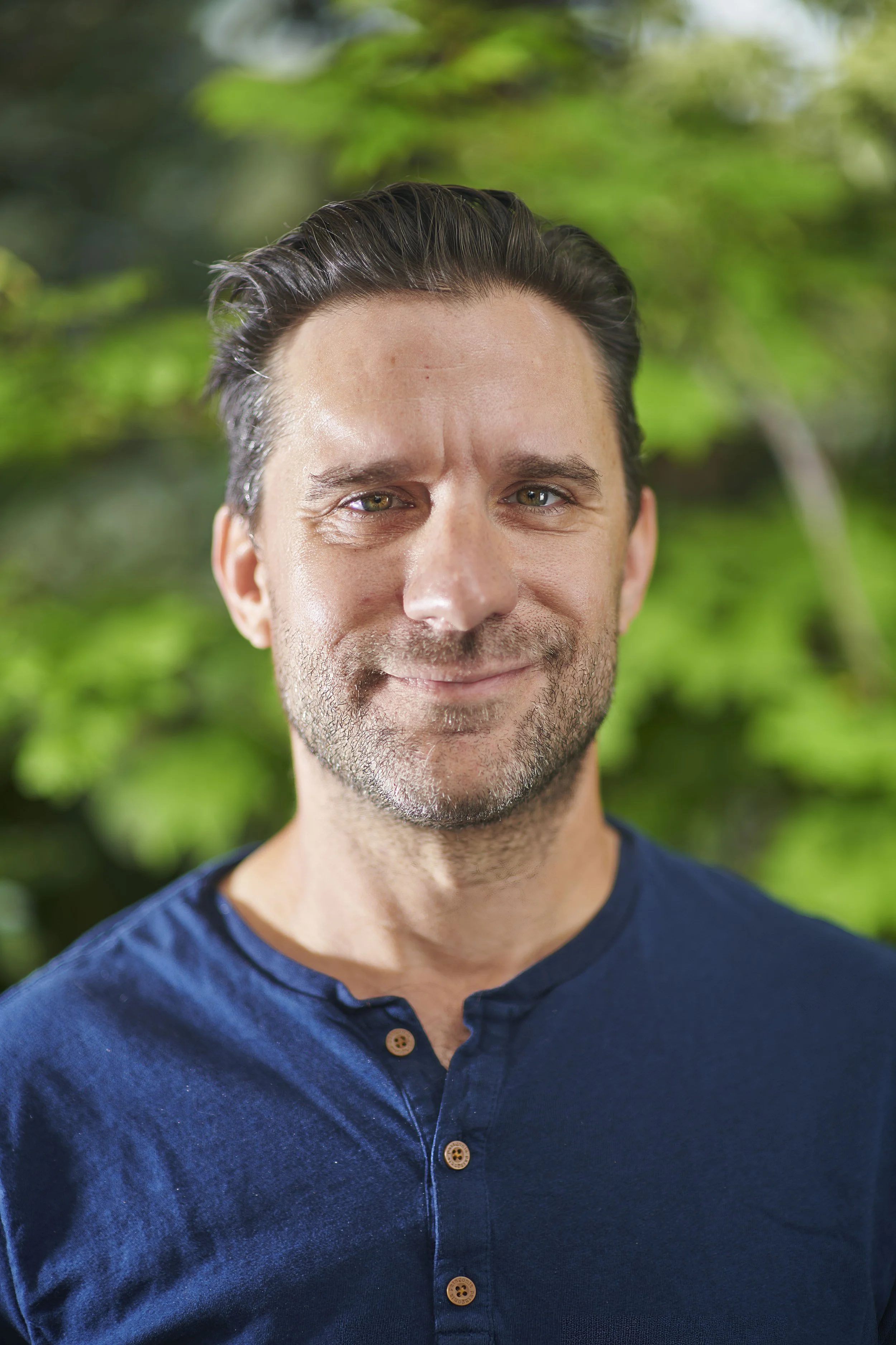 A man with dark hair, slight beard, and green eyes, smiling, wearing a navy blue shirt with wooden buttons, standing outdoors with a blurred green leafy background.