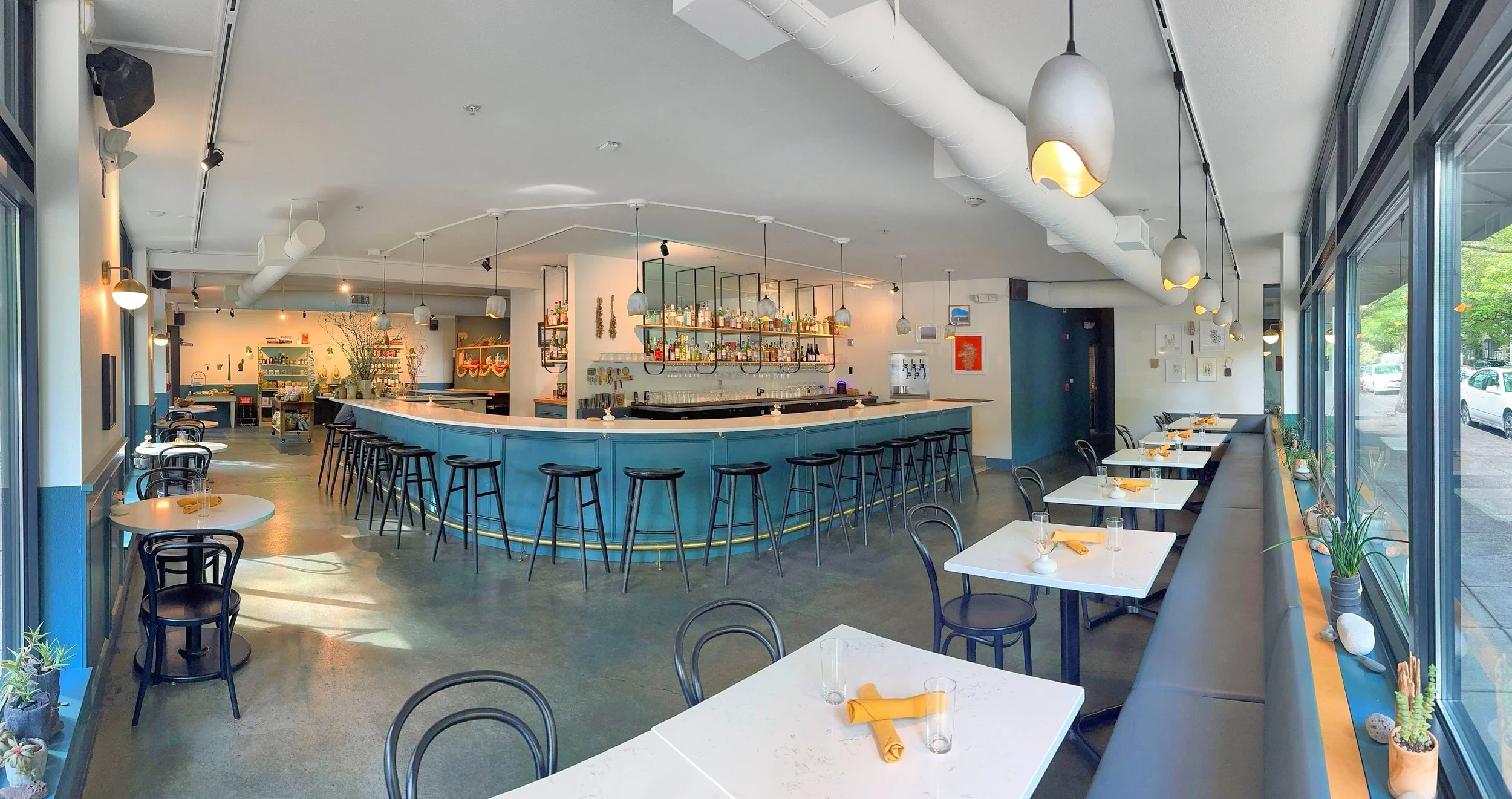 Modern restaurant interior with a bar, white tables set with glasses and yellow napkins, black chairs, large windows with potted plants, and ceiling pendant lights.