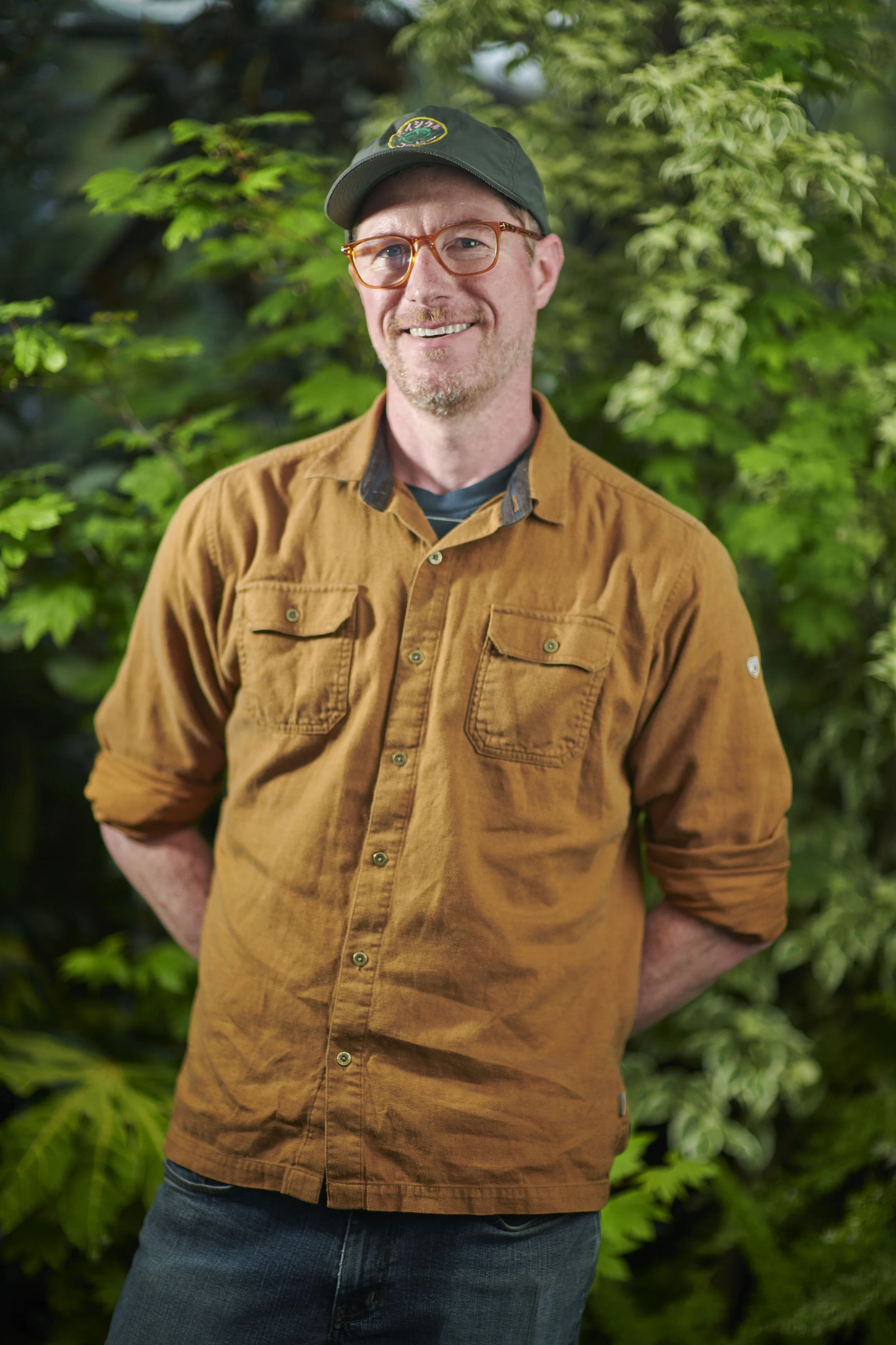 A man wearing glasses, a brown shirt, and a black cap, smiling in front of green foliage.