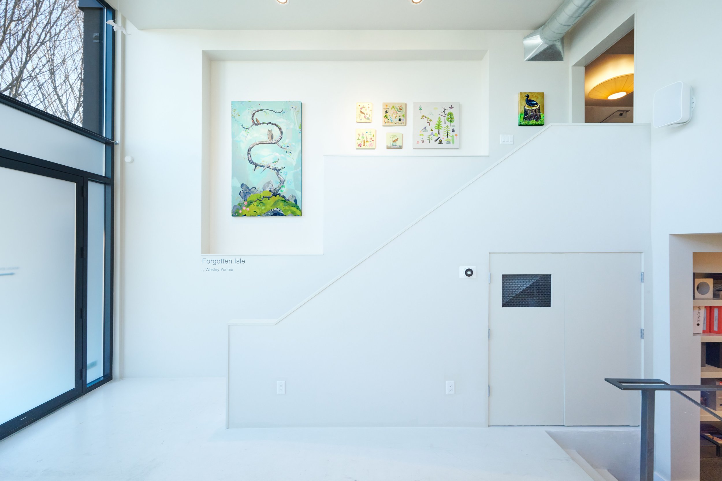 An interior gallery space with white walls and modern design. Artworks are displayed on the wall, including a large vertical painting and several smaller pieces. Large window on the left letting in natural light, and a partial view of a bookshelf on 