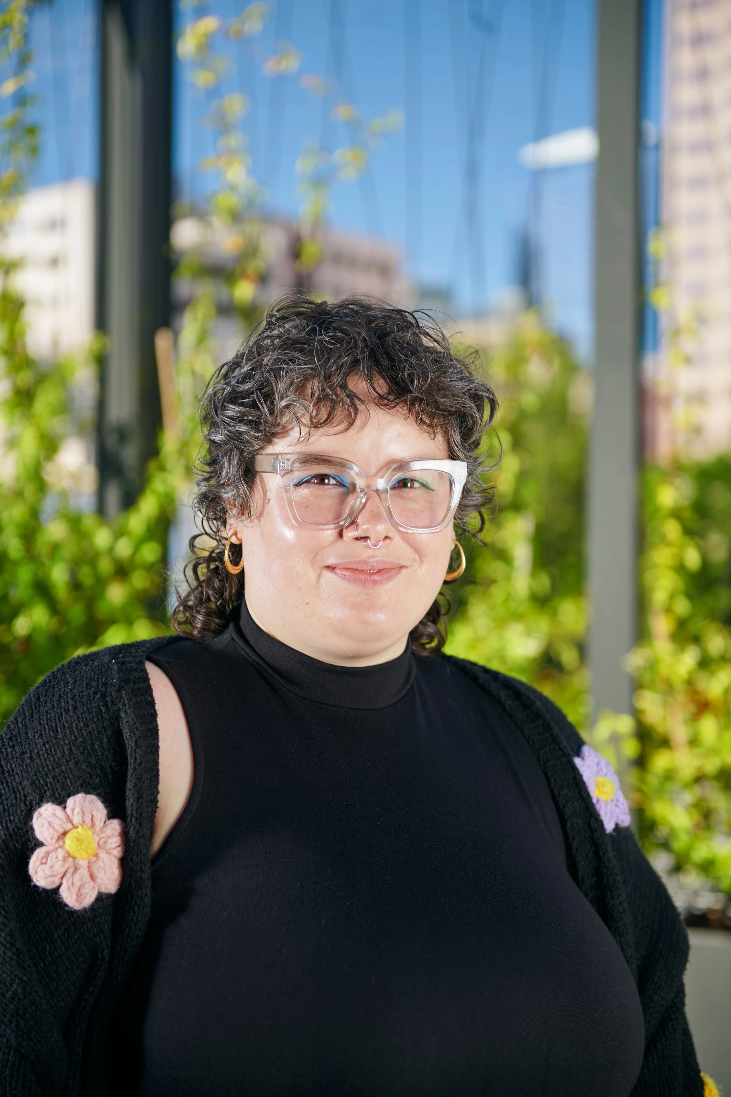 A woman with curly dark hair, clear glasses, and hoop earrings, smiling outdoors with green plants and urban buildings in the background.