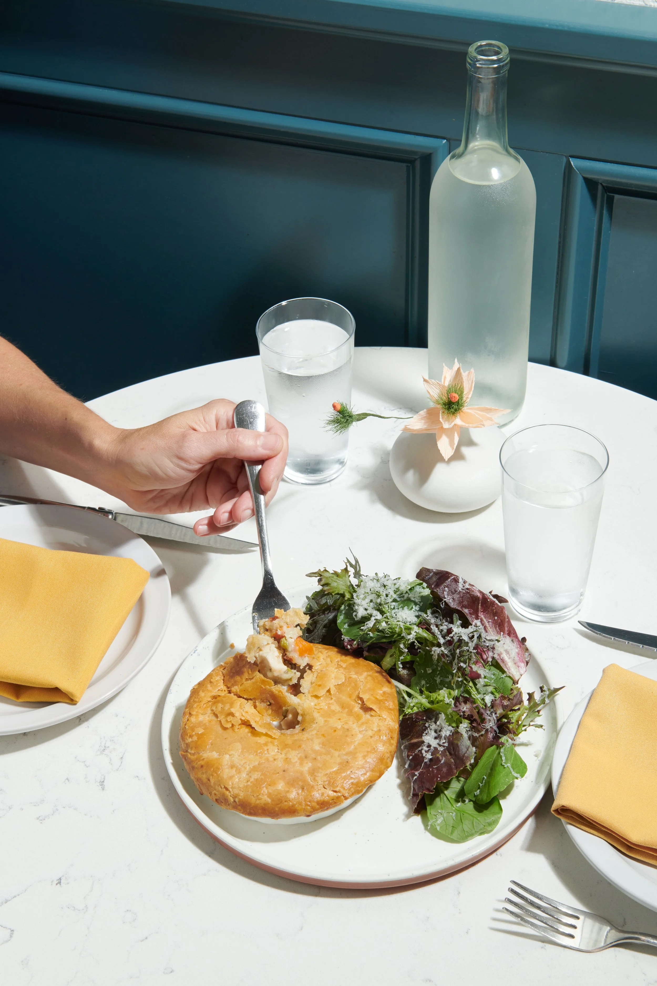 Plate with pie and salad, glasses of water, a vase with a flower, a bottle of water, yellow napkins, and silverware on a white table.