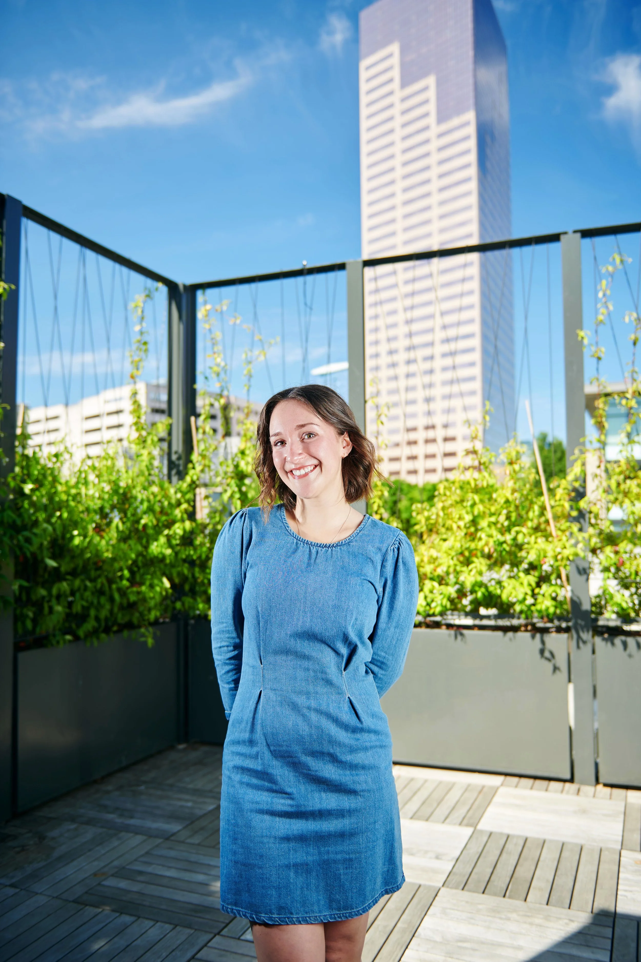 Smiling woman in a denim dress standing on a rooftop garden with a cityscape including a tall building in the background.