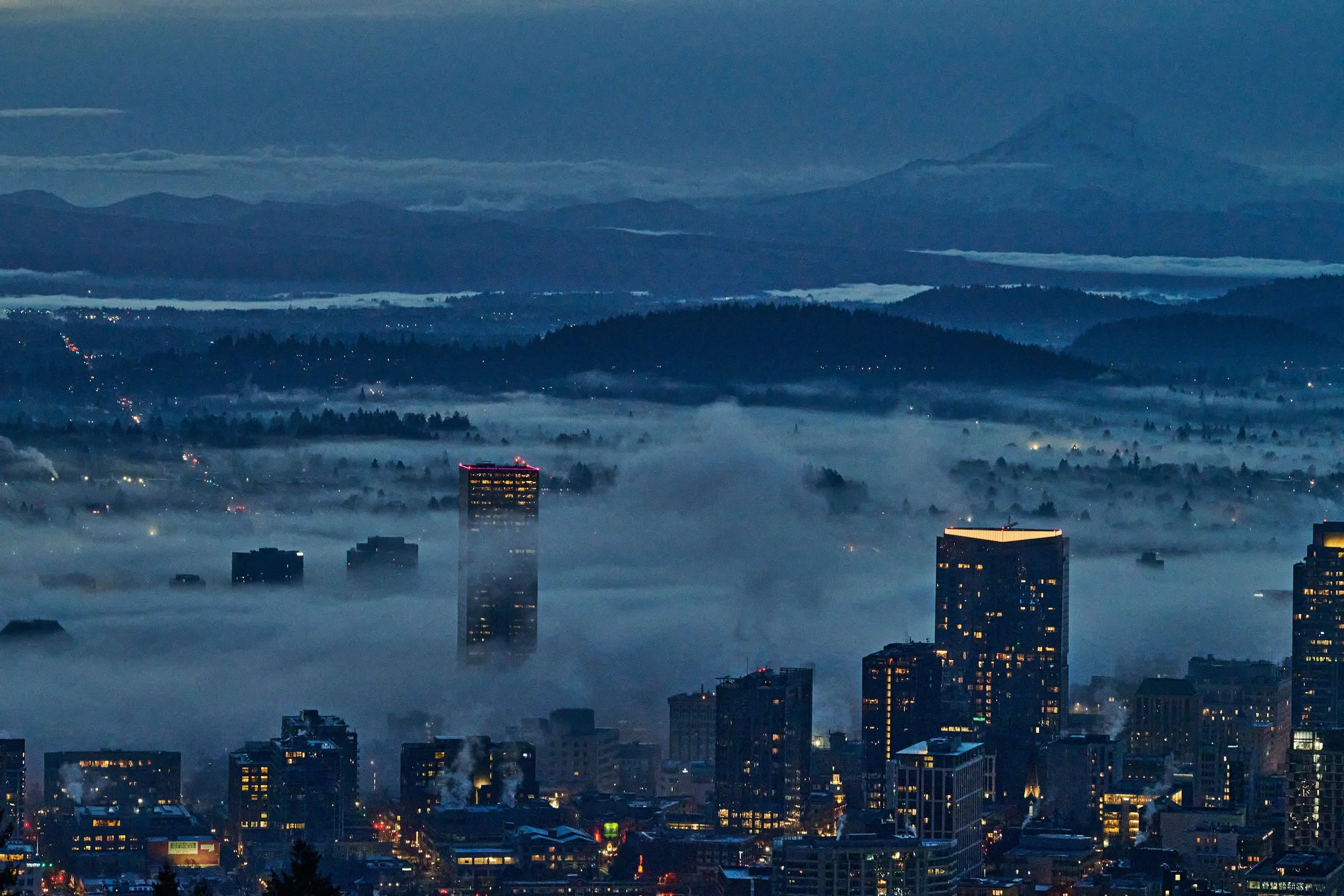 City skyline with tall buildings at dawn, partially covered in fog with hills and mountains in the background.