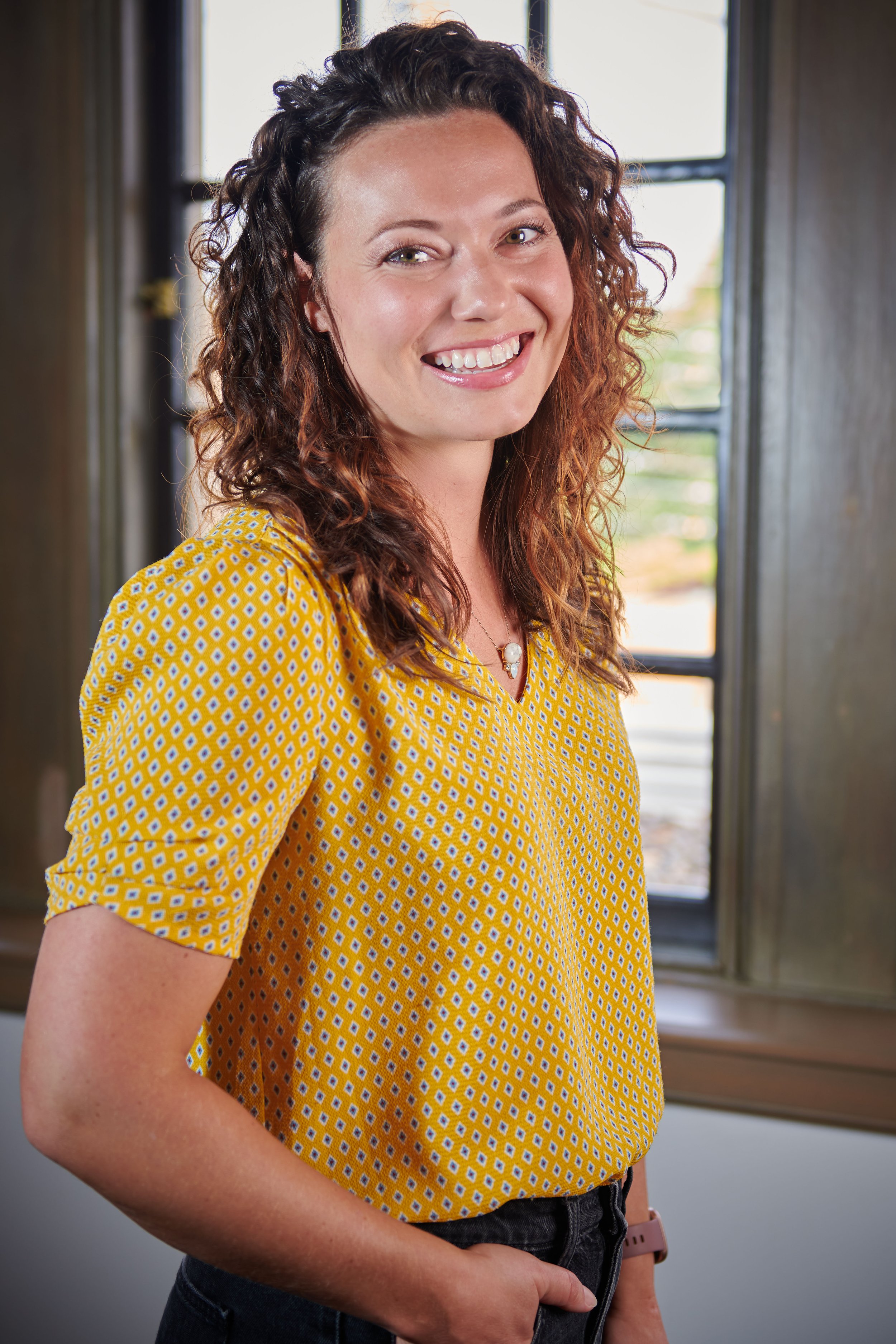 A corporate, environmental portrait of a smiling woman with curly brown hair wearing a yellow patterned blouse standing indoors near window.