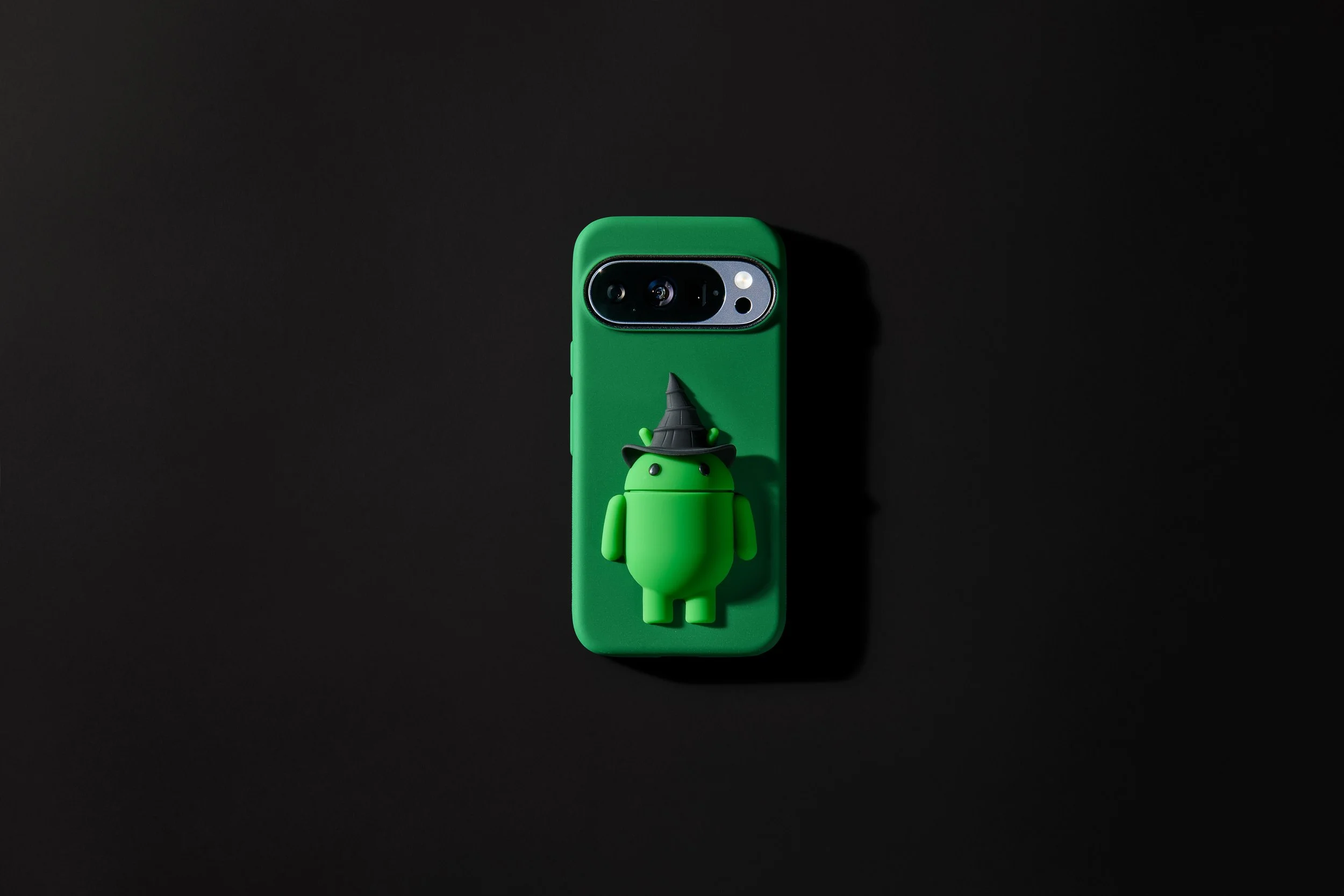 A green smartphone with a 3D Android robot figure wearing a witch's hat on its back, against a black background. The case is merch for the movie Wicked.