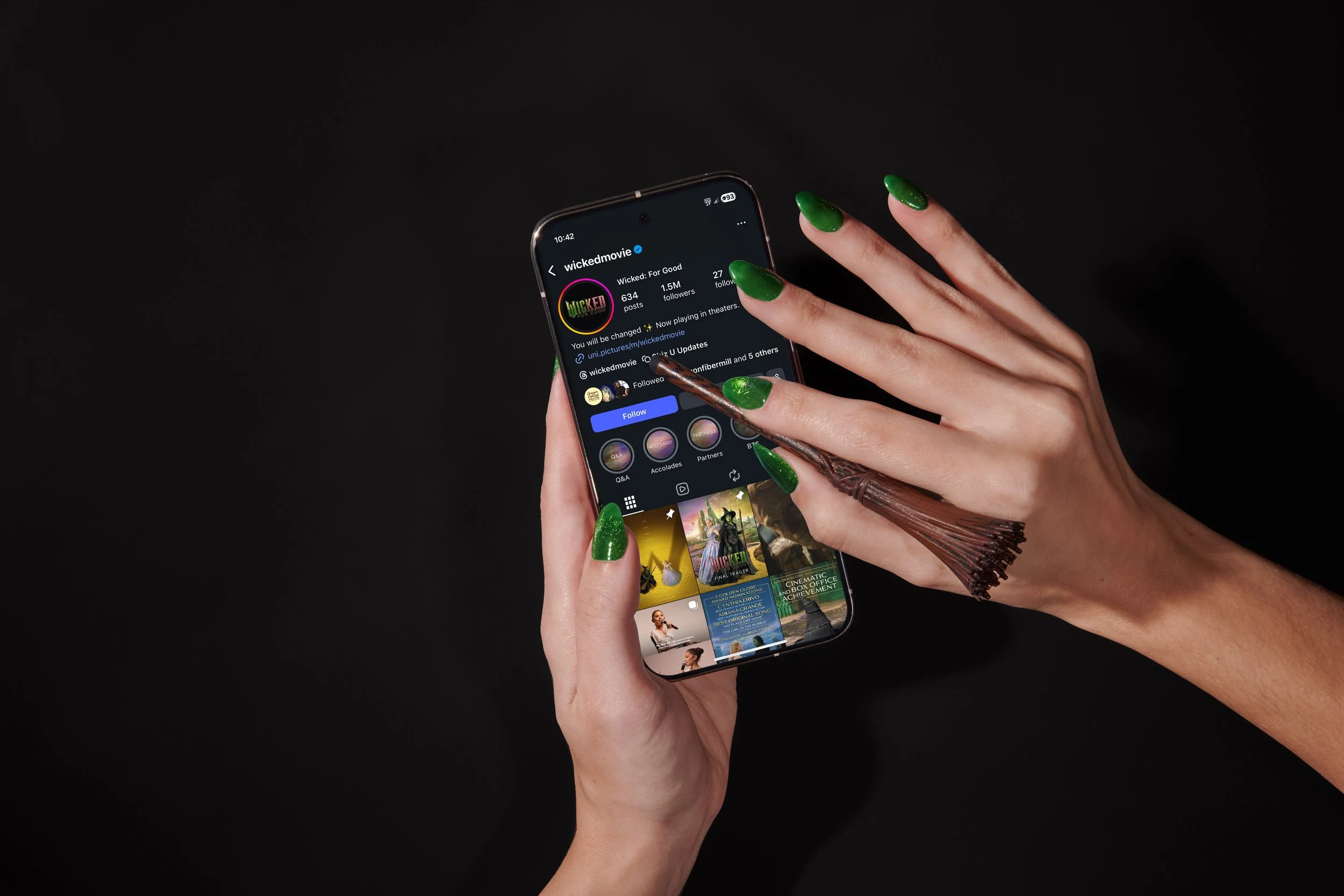 A person with green-painted nails holding a smartphone with a dark background, browsing a social media page related to the movie Wicked on an Android phone.