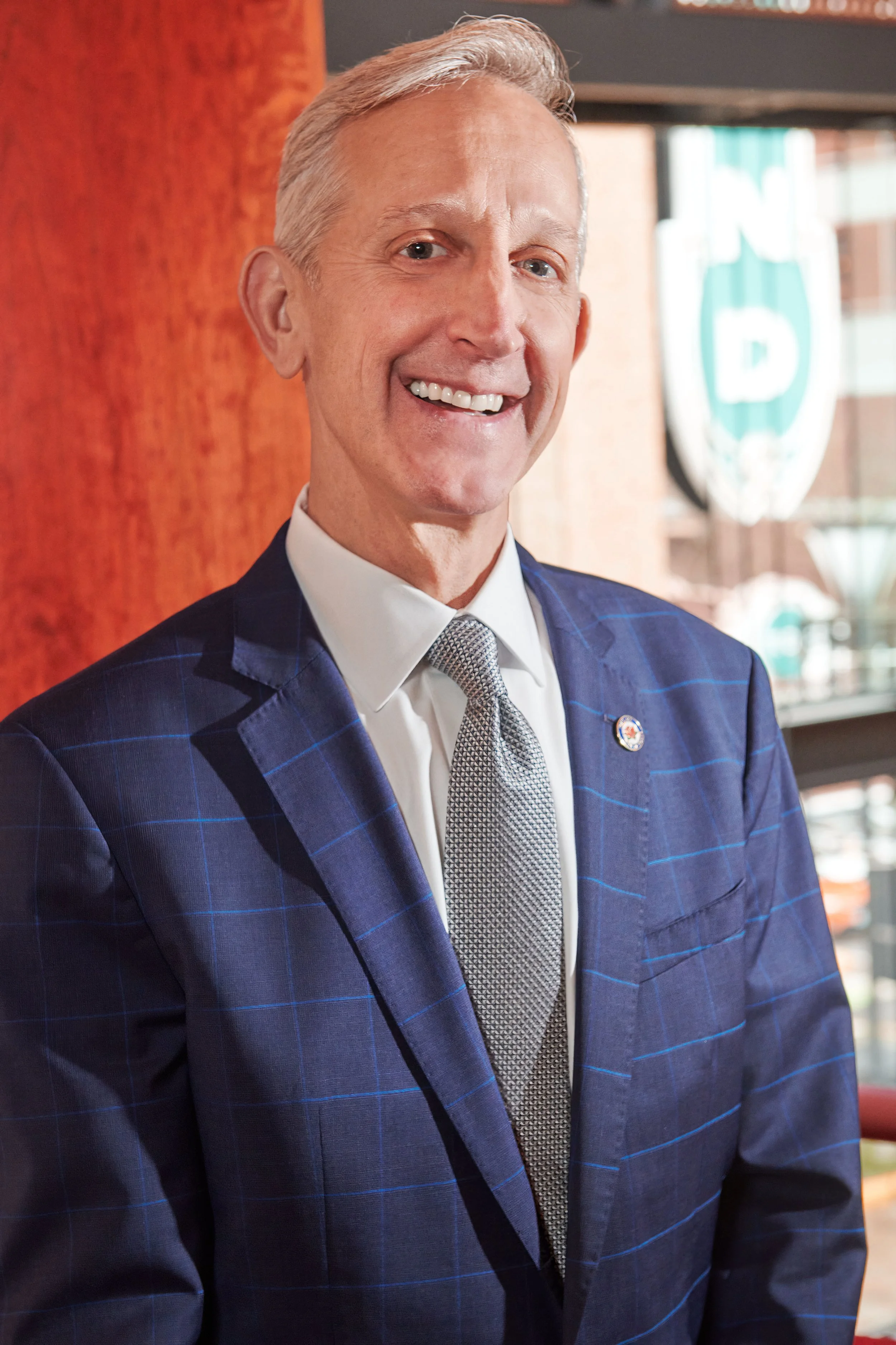 This is a portrait of the mayor of Portland, Oregon Keith Wilson. This is a polished portrait of a smiling man with gray hair in a checkered blue suit and a patterned tie, standing indoors near a window with a blurred Portland sign in the background.