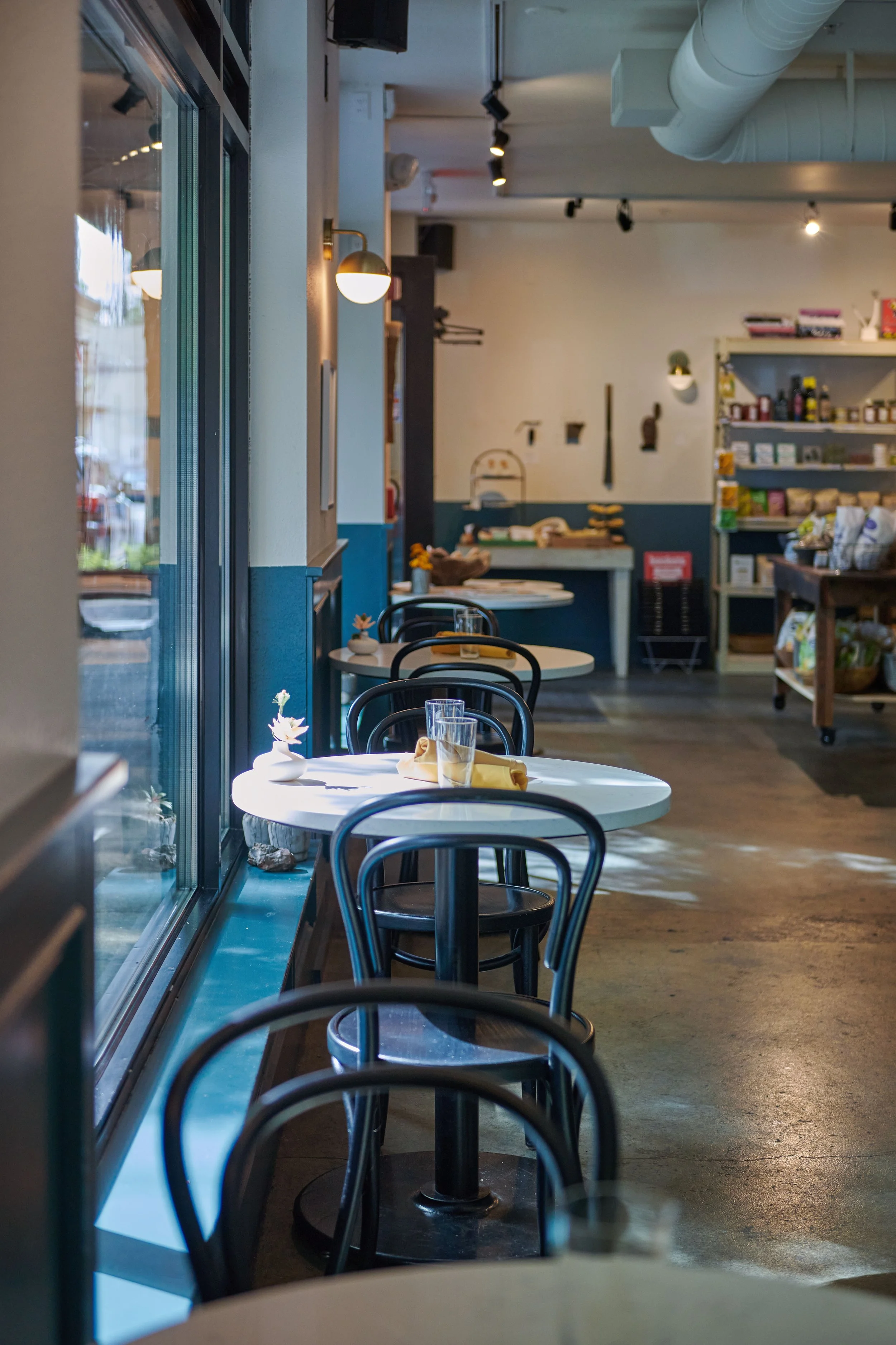 Empty cafe with round tables and black chairs, sunlight coming through large windows, shelves with assorted food and products in the background.