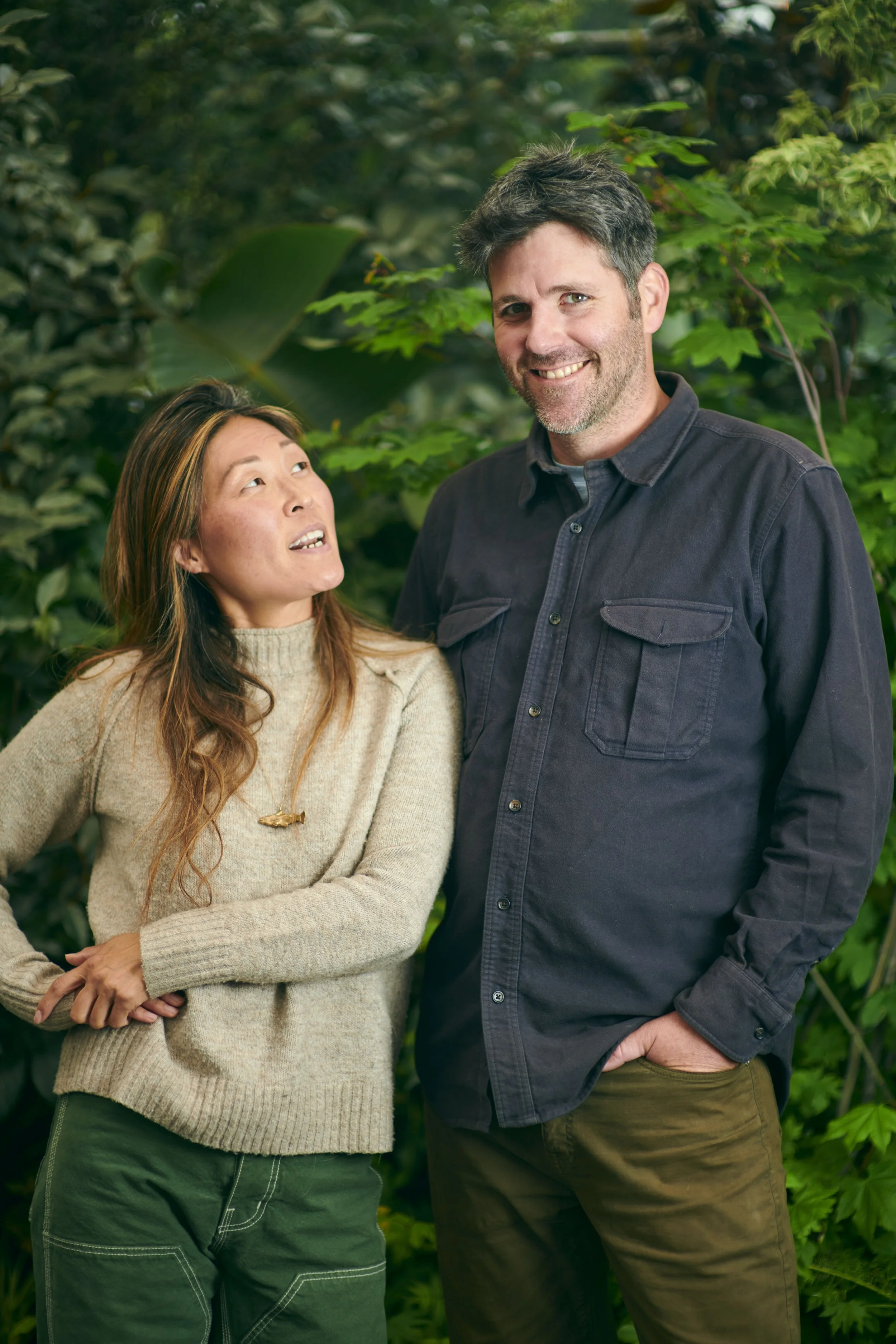 A woman and a man standing outdoors among green foliage, engaging in conversation.