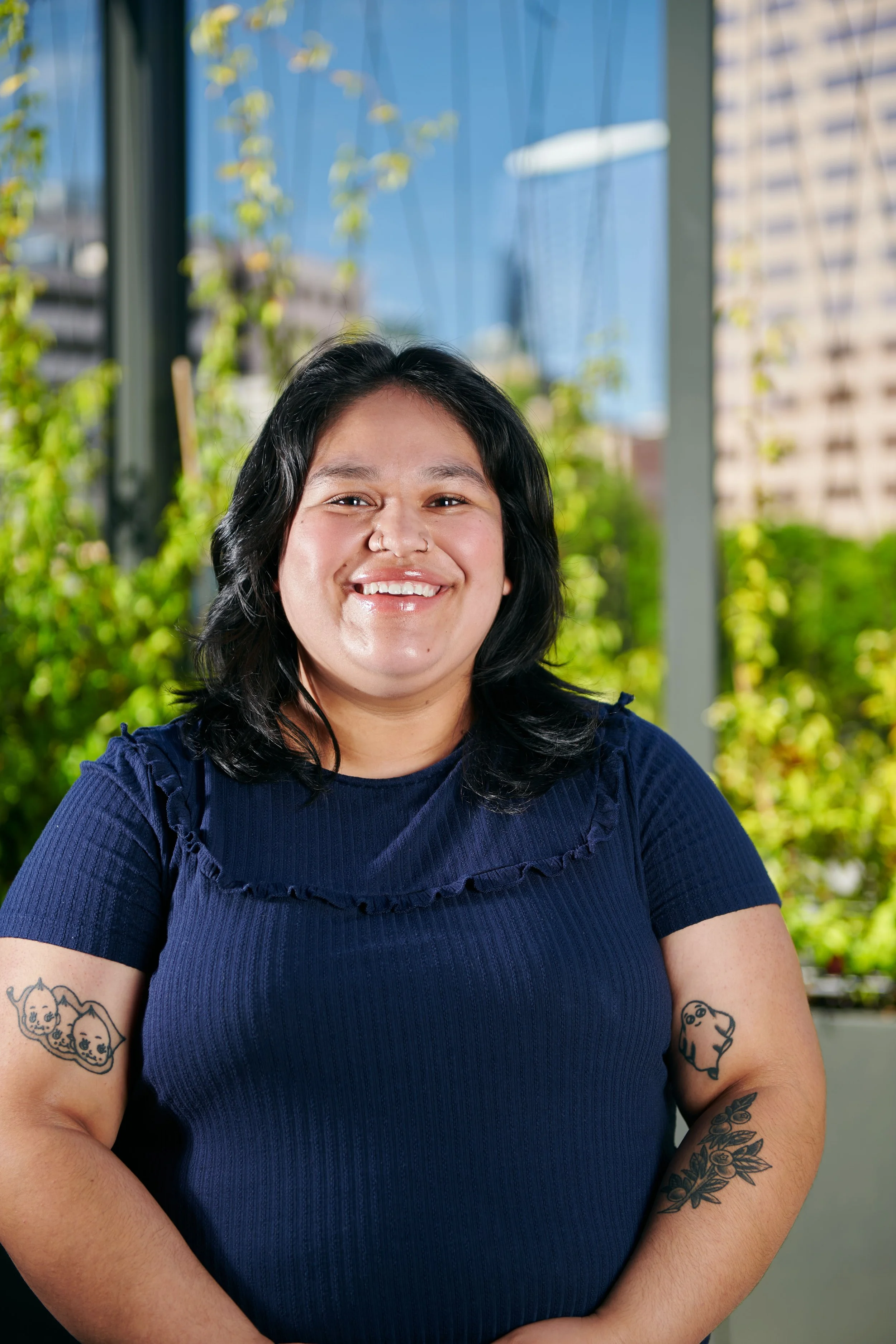A woman with black hair and tattoos on her arms, smiling in front of a lush green background with city buildings and blue sky visible.