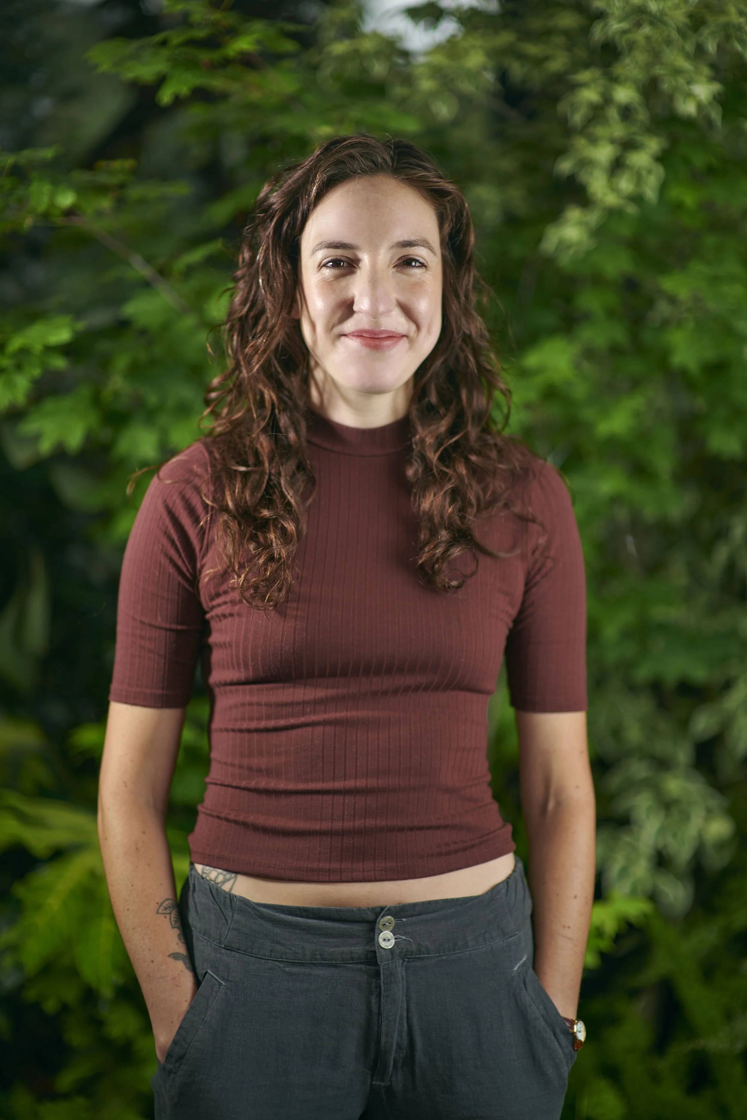 A young woman with curly brown hair, wearing a maroon ribbed shirt and dark pants, standing outdoors in front of green foliage.