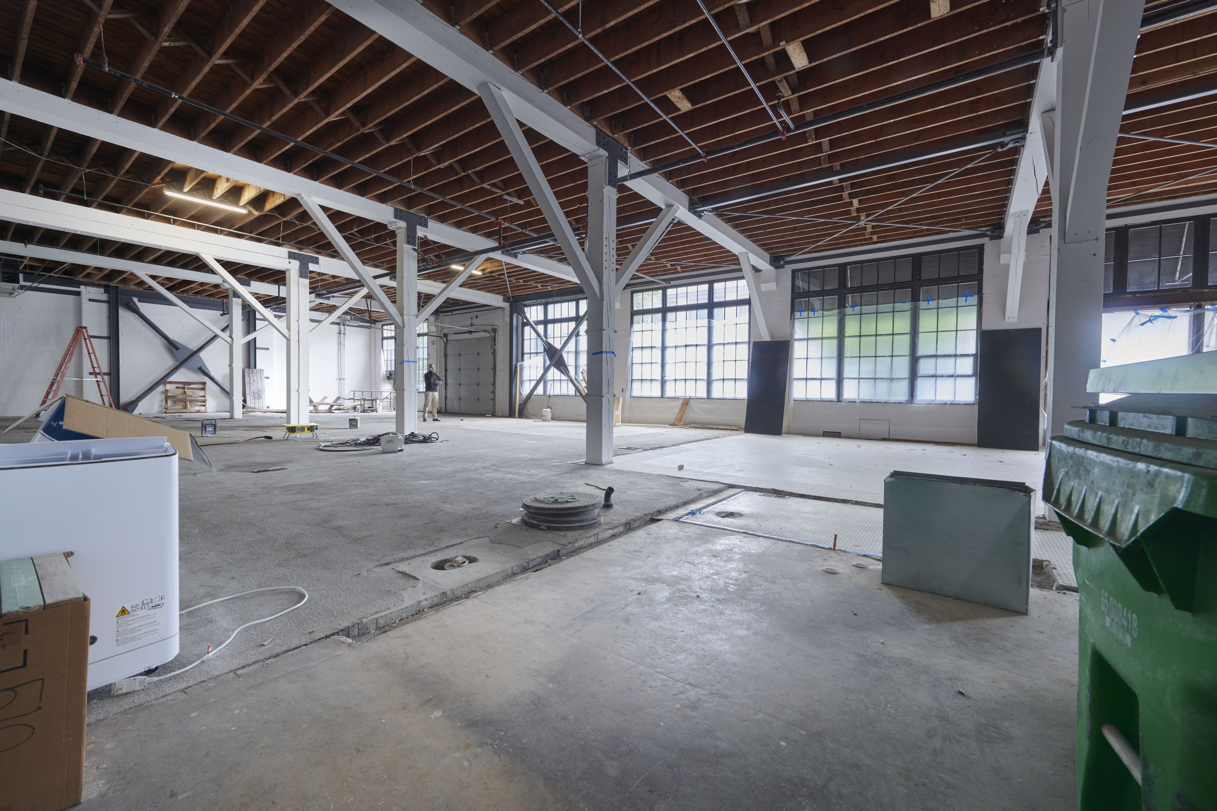 Interior of a building under construction with exposed ceiling beams, large windows, construction tools, and equipment scattered on the floor.