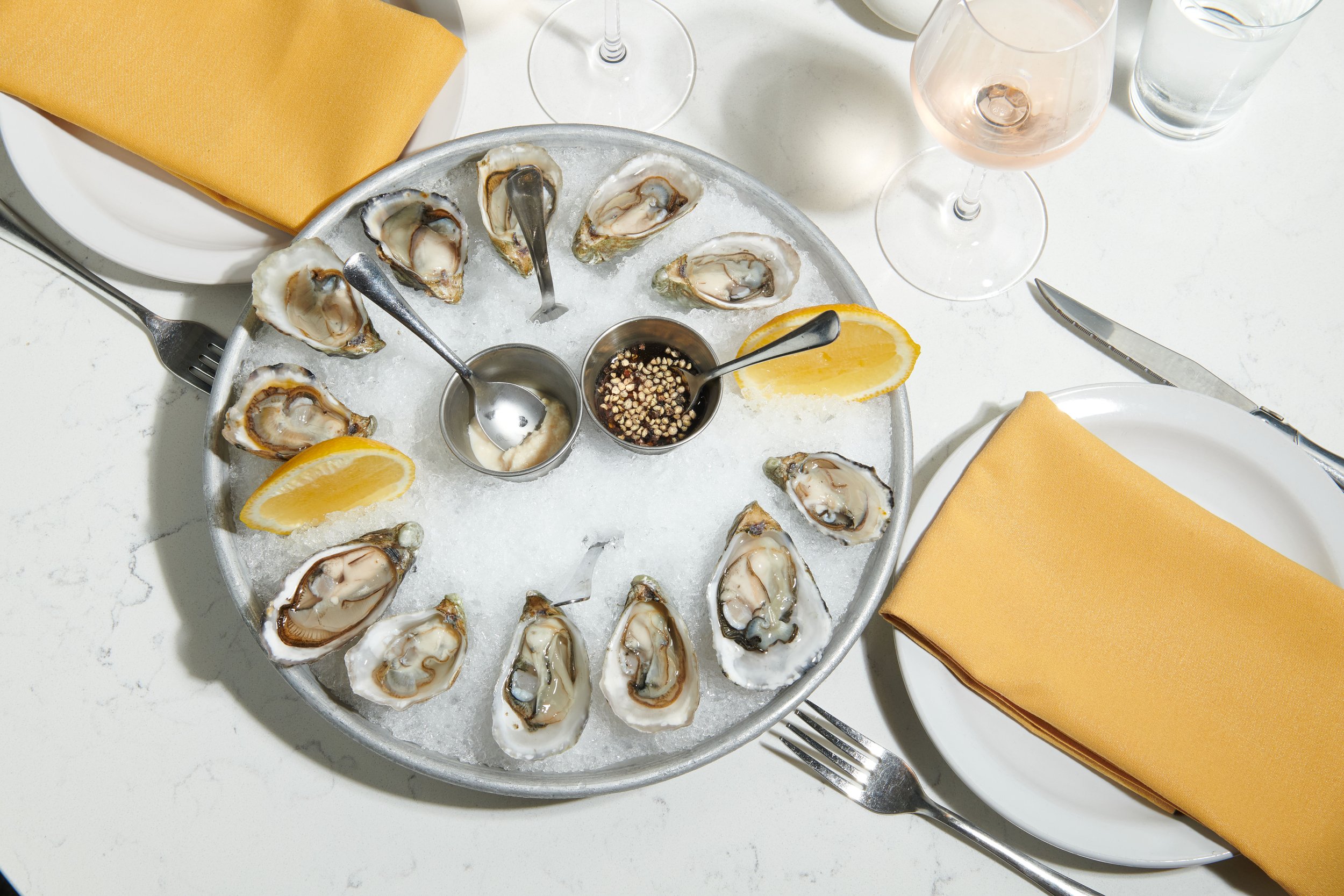 A seafood dinner featuring raw oysters on a bed of ice, lemon wedges, and condiments, set at a table with yellow napkins and glassware.