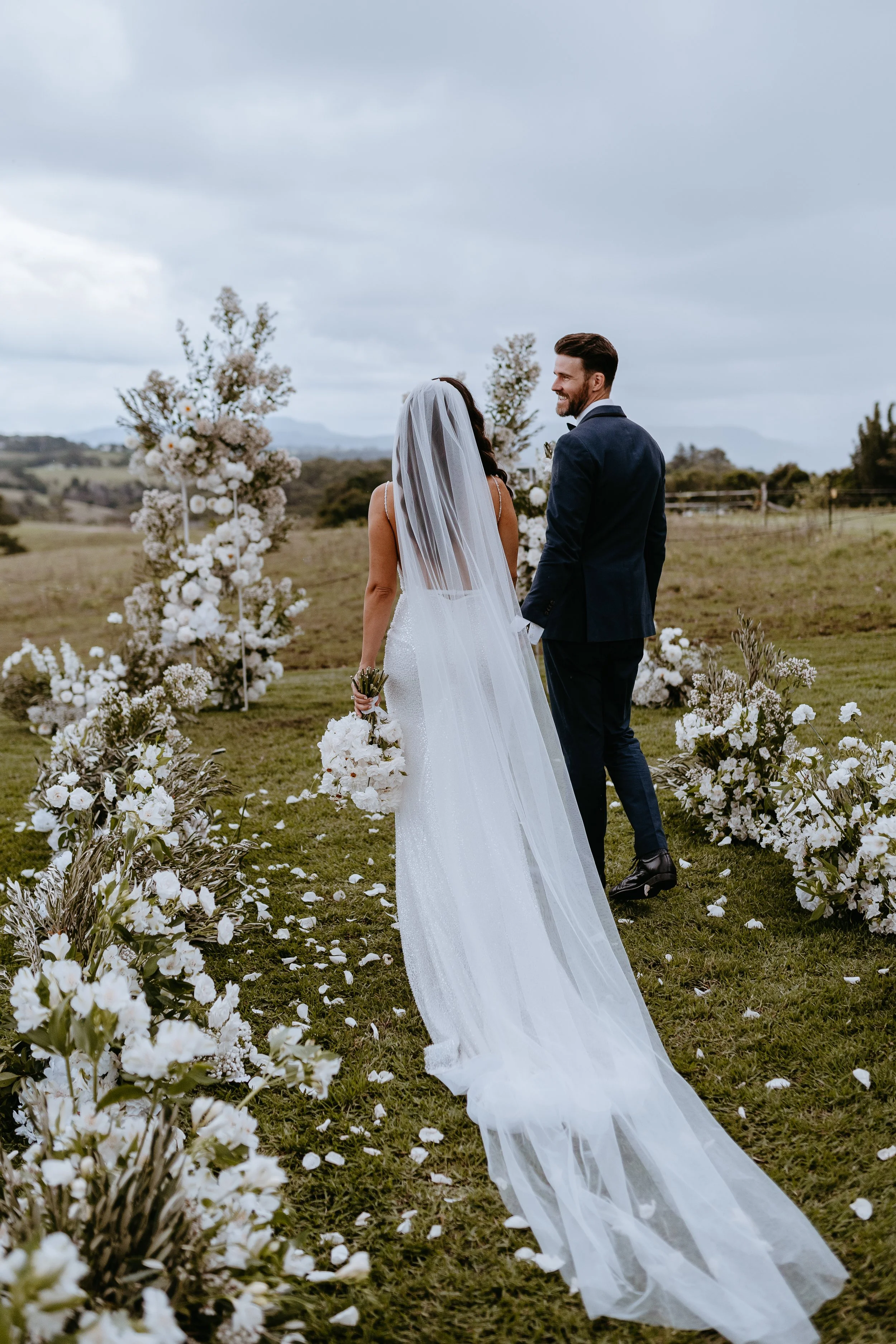 A bride and groom holding hands during their outdoor wedding ceremony on a cloudy day, surrounded by floral arrangements with white flowers and greenery.