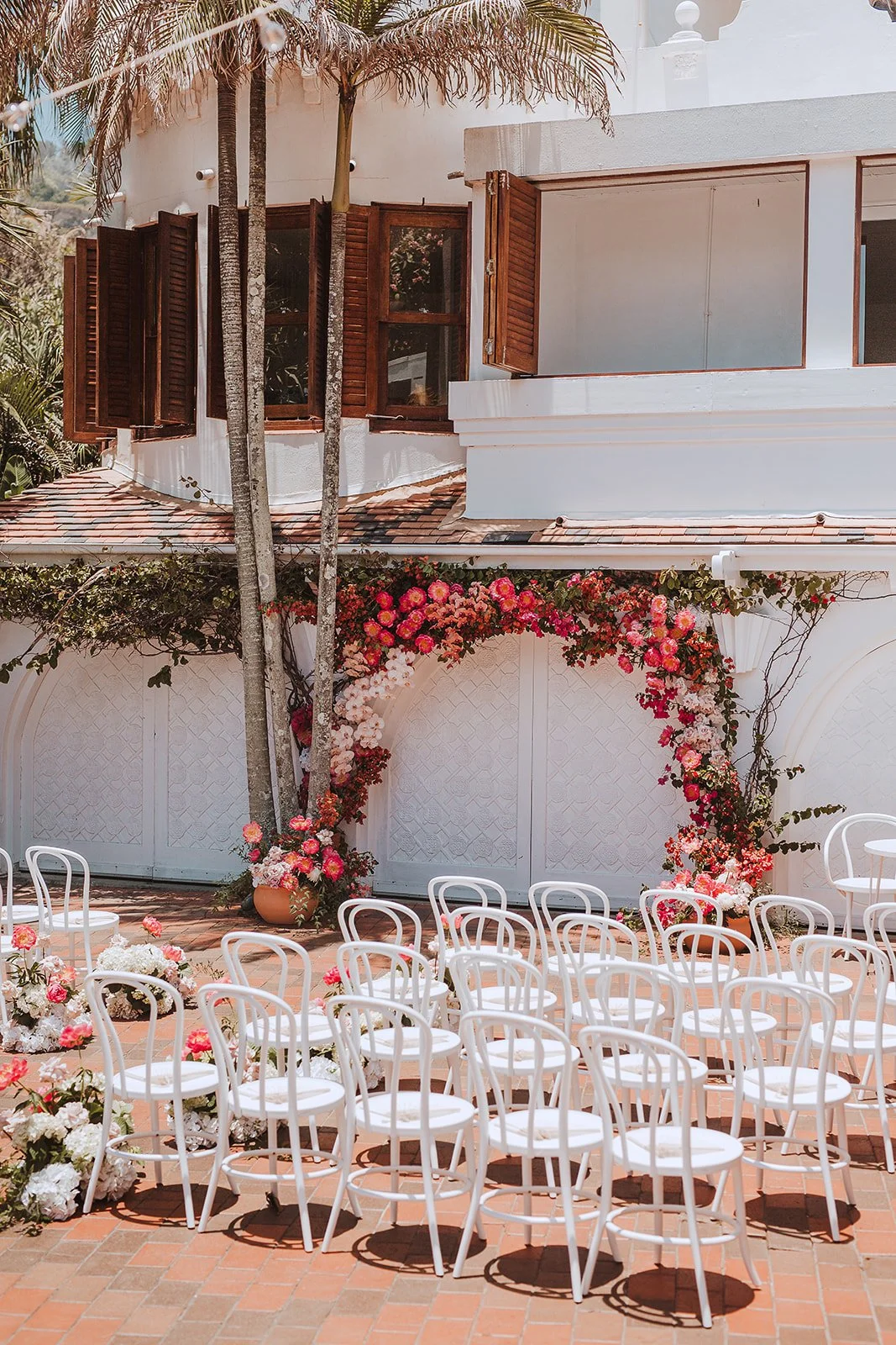 Outdoor wedding setup with white chairs arranged in front of a floral arch and a white building with wooden shutters.