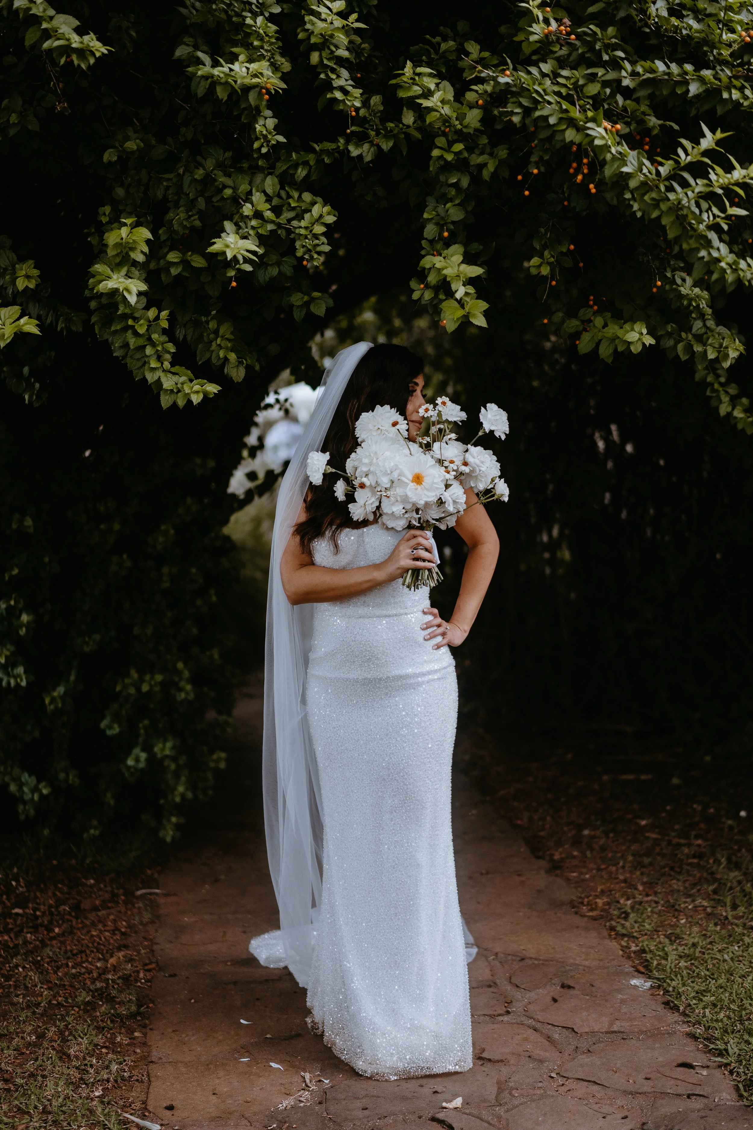 A bride in a white, glittery wedding dress stands on a stone pathway under a green arch of leafy branches. She holds a bouquet of white daisies and large white flowers, with her right hand on her hip, and wears a veil.