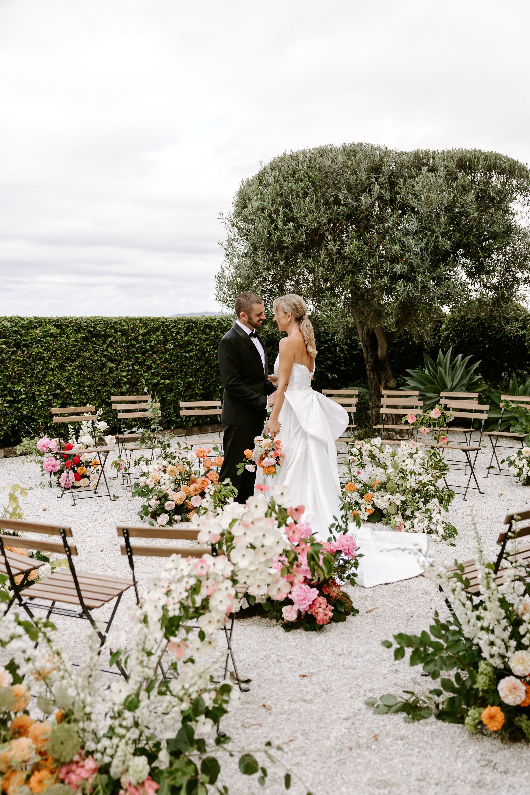 Bride and groom standing together at an outdoor wedding ceremony, surrounded by colorful flowers and greenery.