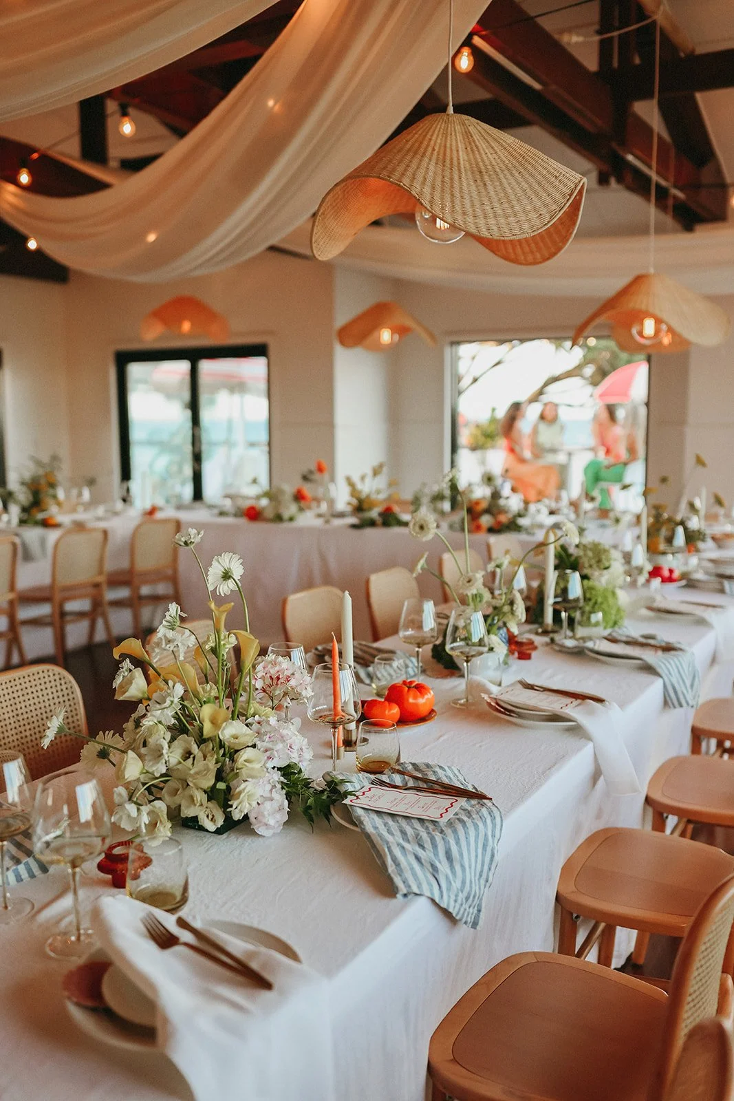 A decorated banquet table with floral centerpieces, candles, glasses, and place settings in a well-lit indoor space with hanging woven light fixtures.