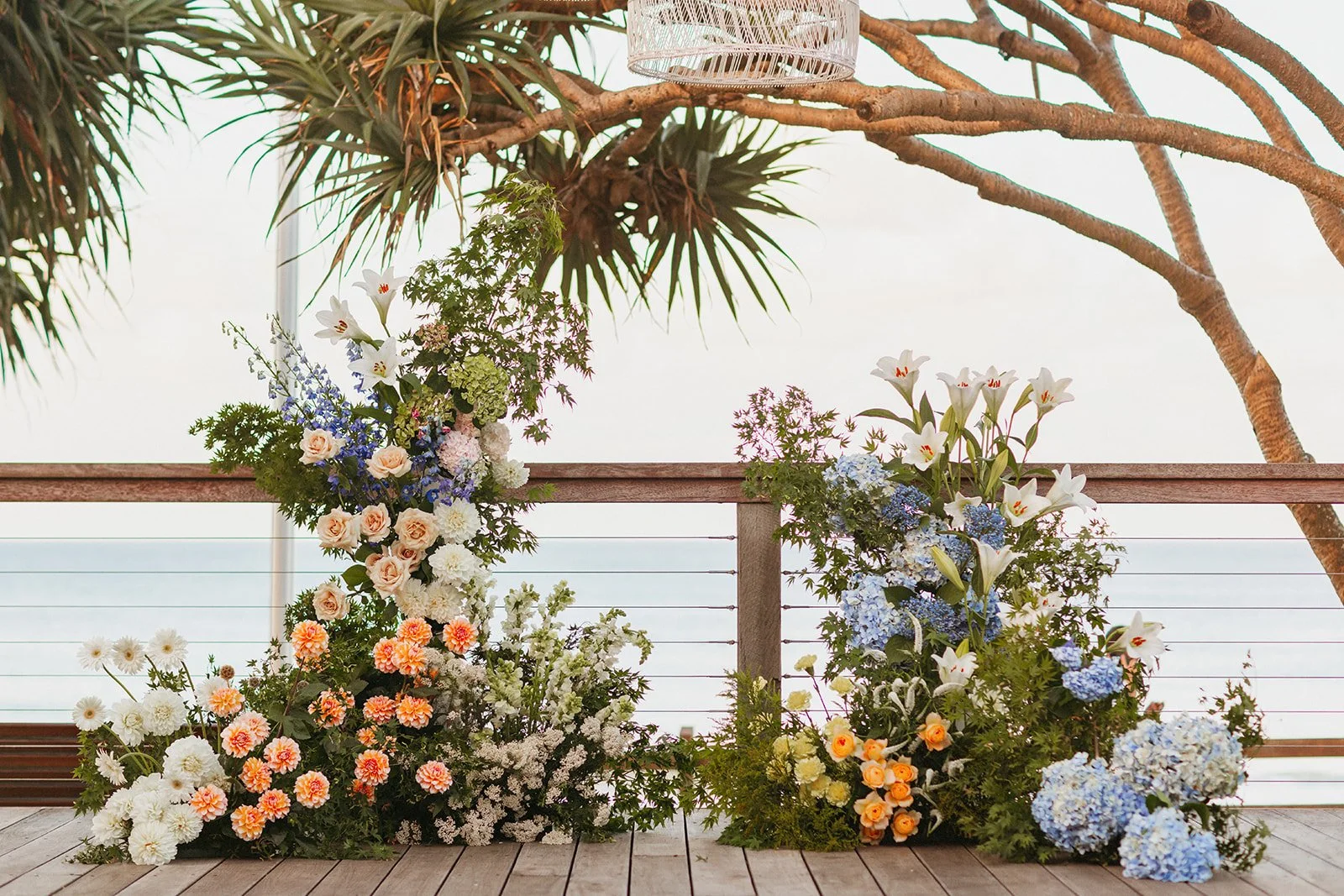 Colorful floral arrangements with white lilies, blue hydrangeas, pale pink roses, orange roses, and white chrysanthemums on a wooden deck with a railing and a large tropical tree