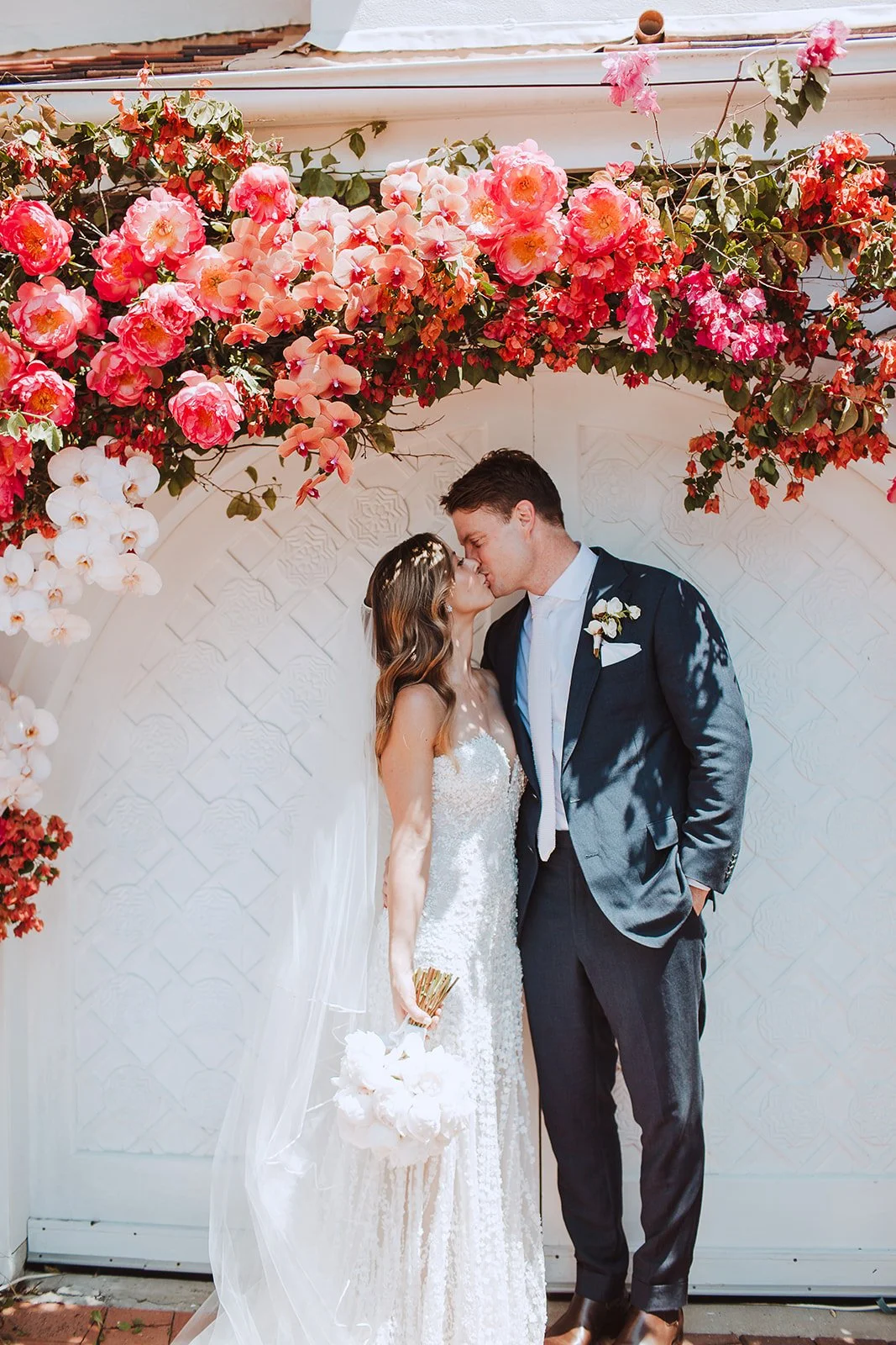 Bride and groom sharing a kiss under a floral arch with pink, red, and white flowers at a wedding.