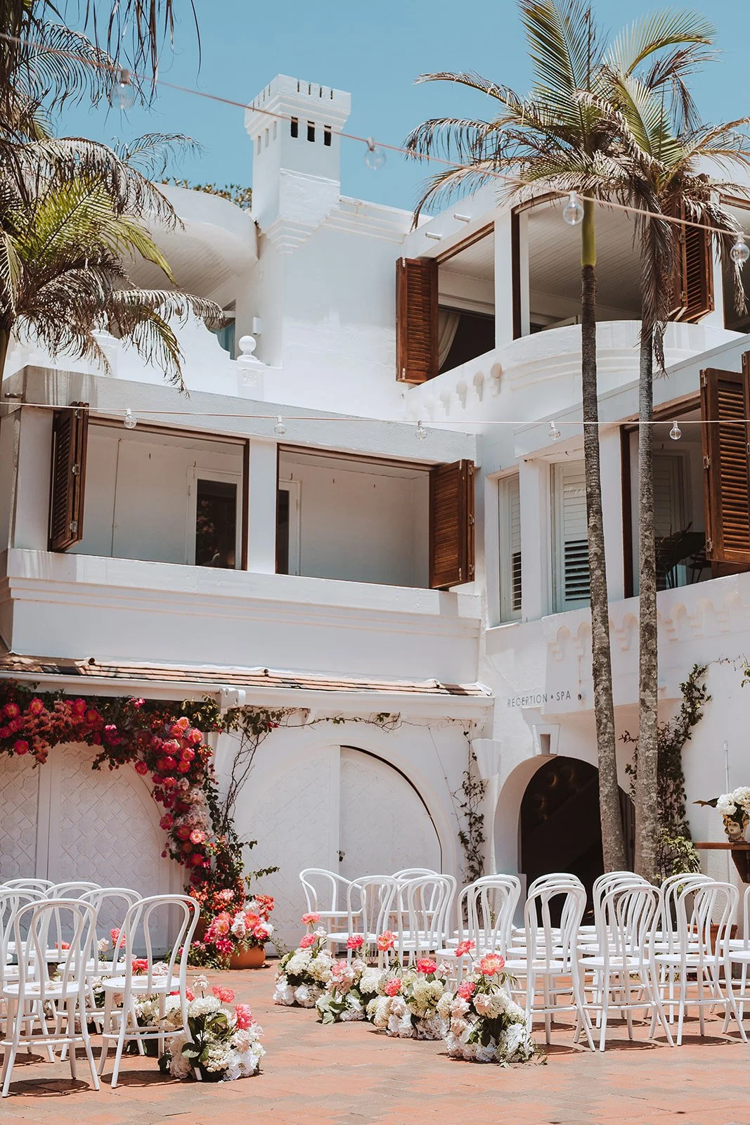 A white building with open wooden shutters, balconies, and palm trees. An outdoor ceremony setup with white chairs and floral arrangements, and string lights overhead.