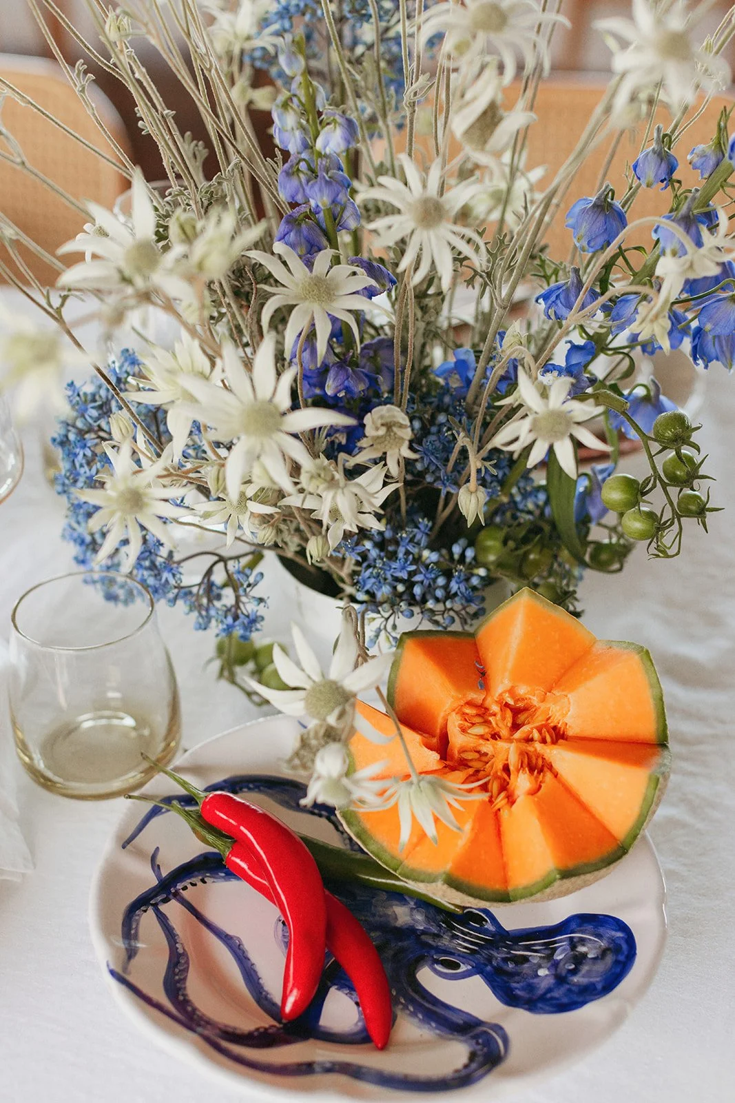 A table with a large bouquet of white and blue flowers, a half-cut orange melon, red chili peppers, and a clear glass of white wine on a white tablecloth.
