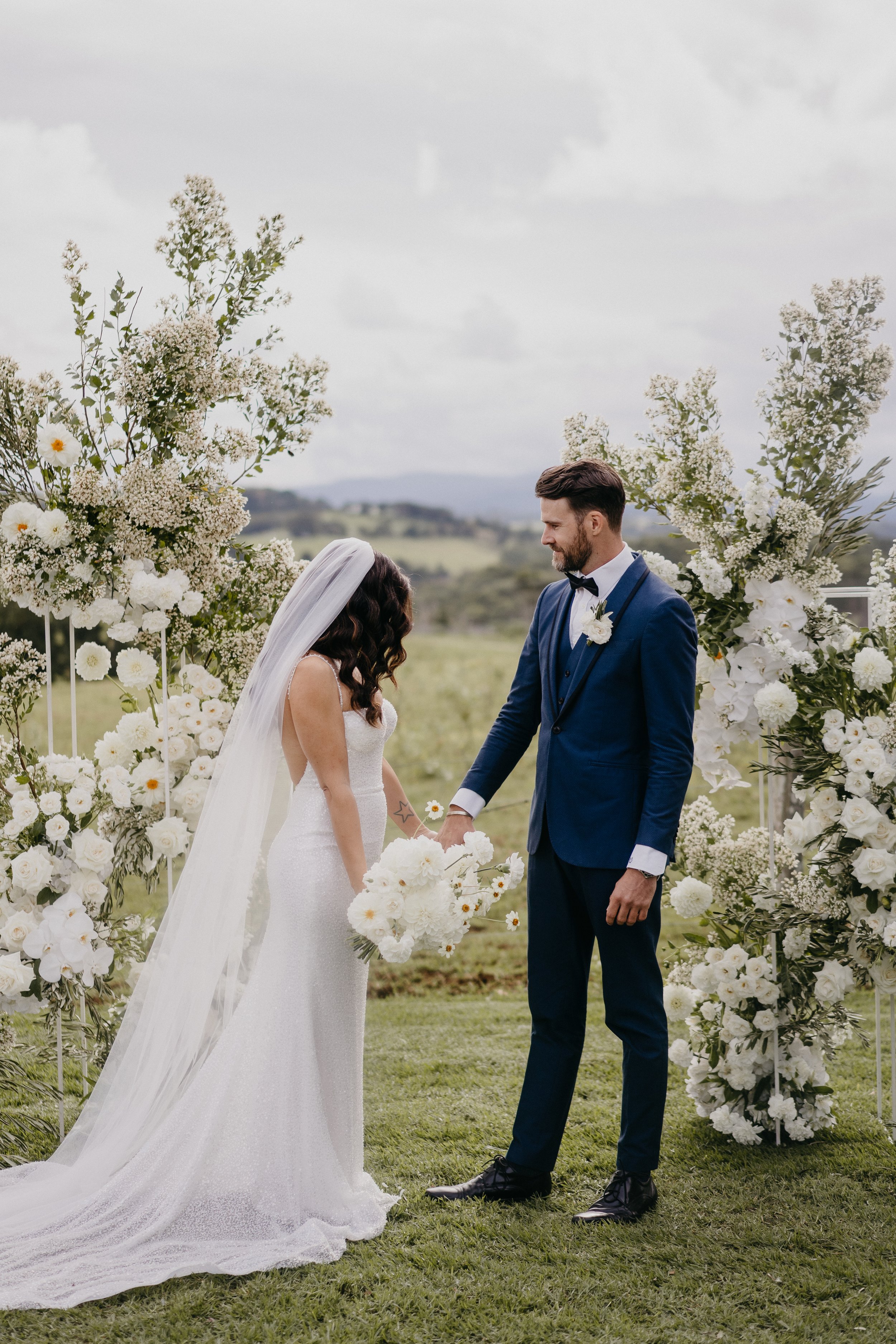 Bride and groom holding hands during outdoor wedding ceremony surrounded by white floral arrangements