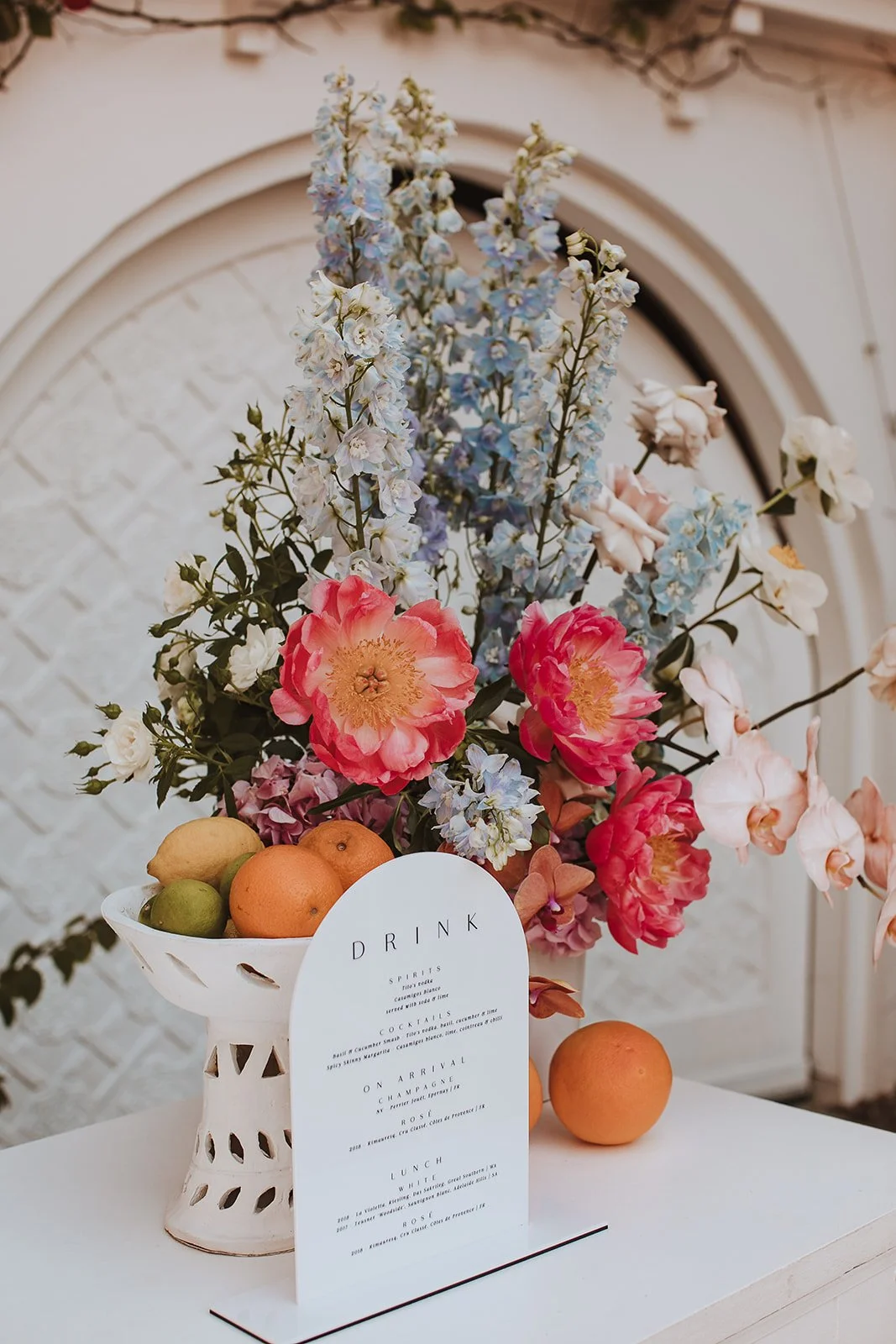 A floral arrangement with pink, white, and blue flowers, a white decorative fruit bowl containing lemons, limes, and oranges, and a white menu card on a white table against a white textured wall.