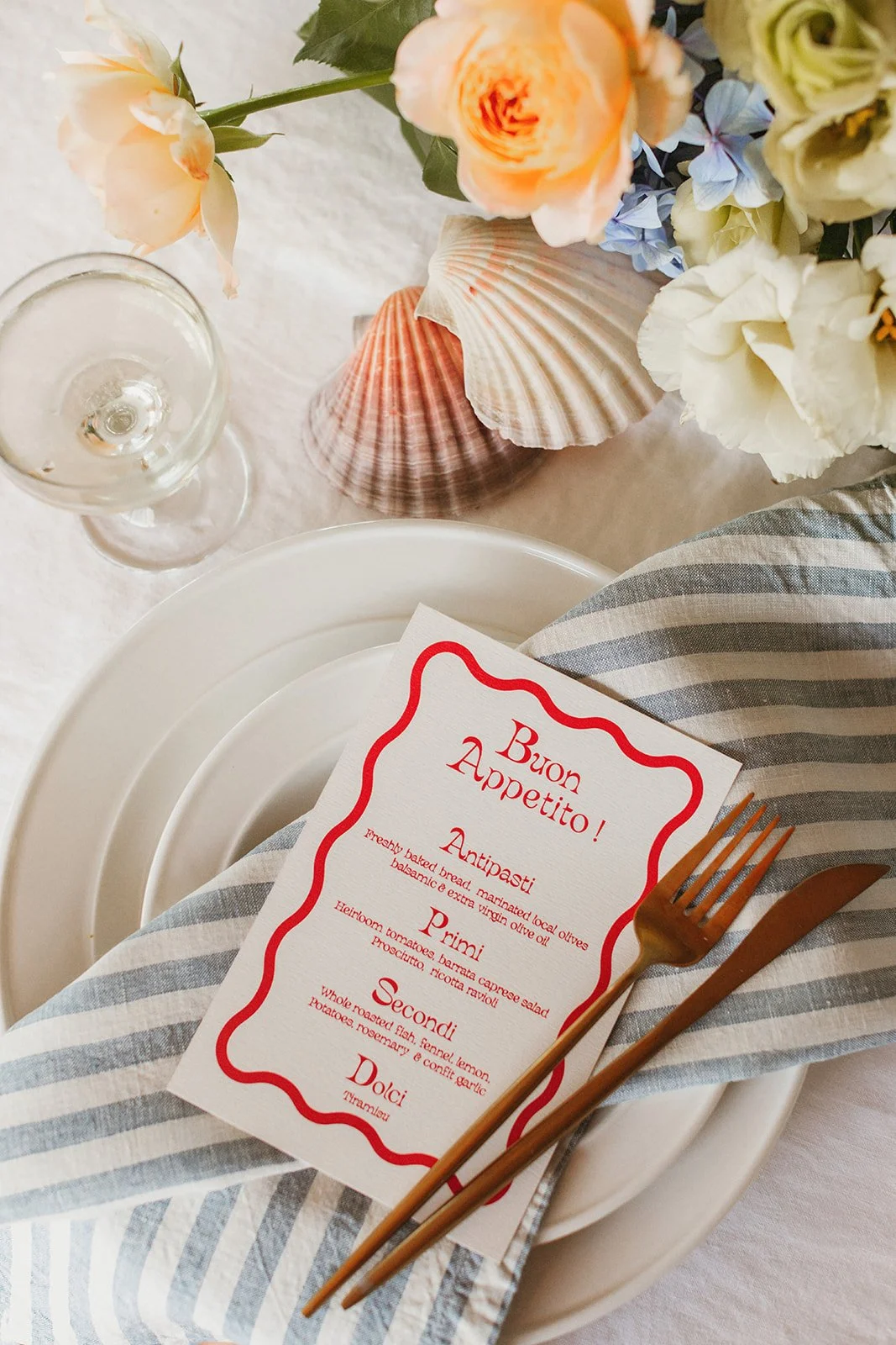 Table setting with a white plate, striped napkin, gold fork and knife, a menu card with red text, a glass of white wine, a flower arrangement with roses, hydrangeas, and other flowers, and seashells on a white tablecloth.