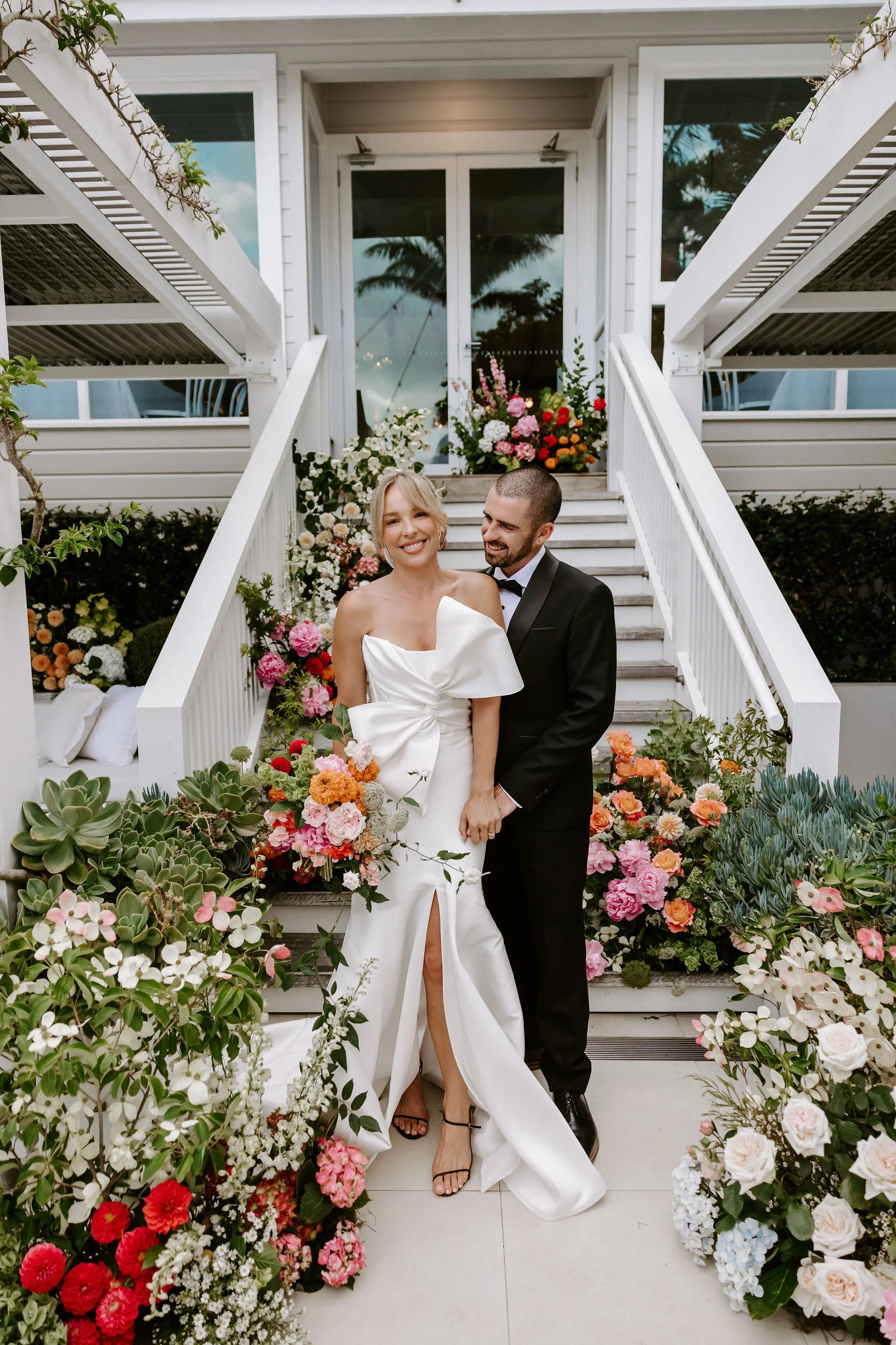 A newly married couple standing on stairs decorated with colorful flowers, with a woman in a white gown holding a bouquet and a man in a black tuxedo, smiling and embracing.