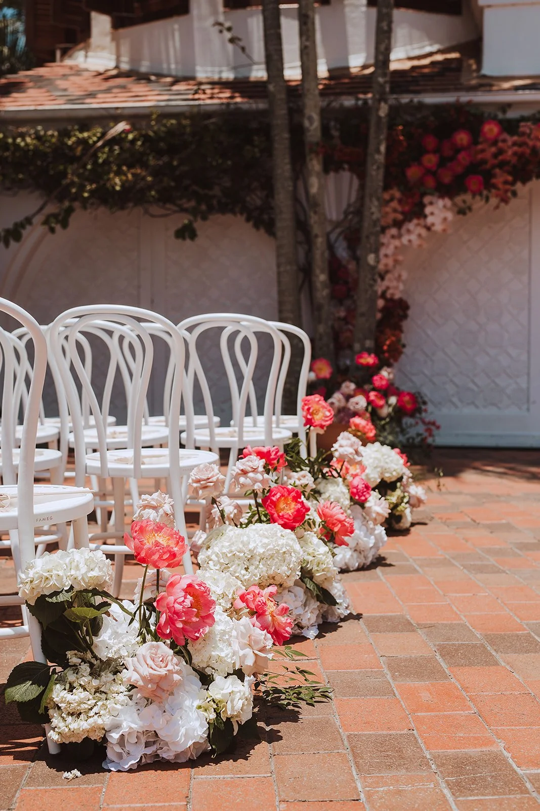 White chairs arranged outdoors with pink, white, and cream flower arrangements along the edge of a tiled patio, with a backdrop of trees and a decorative wall with flowers.