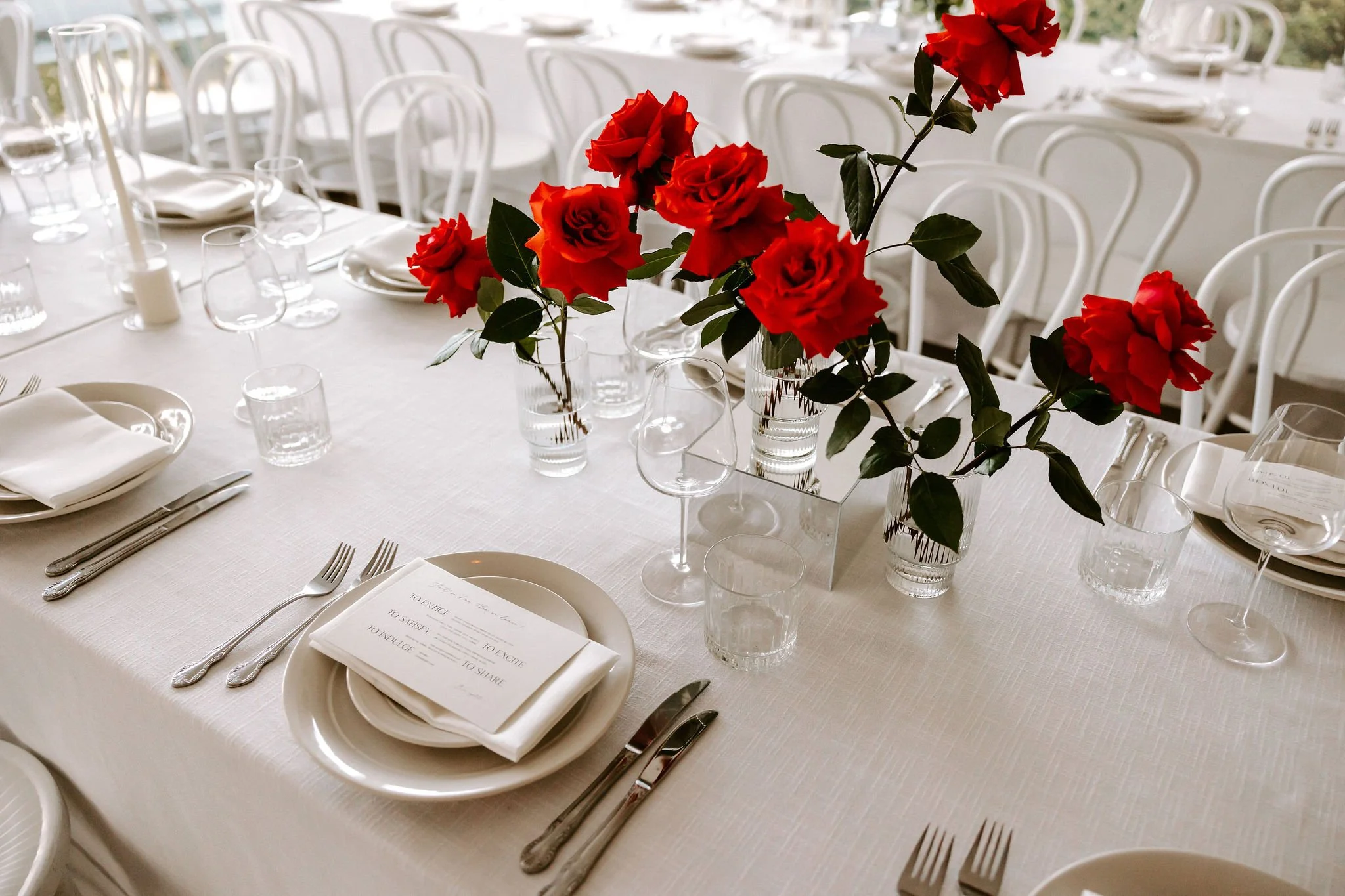 Elegant dining table set with white tablecloth, white plates, silverware, glassware, and a centerpiece of red roses in clear vases.