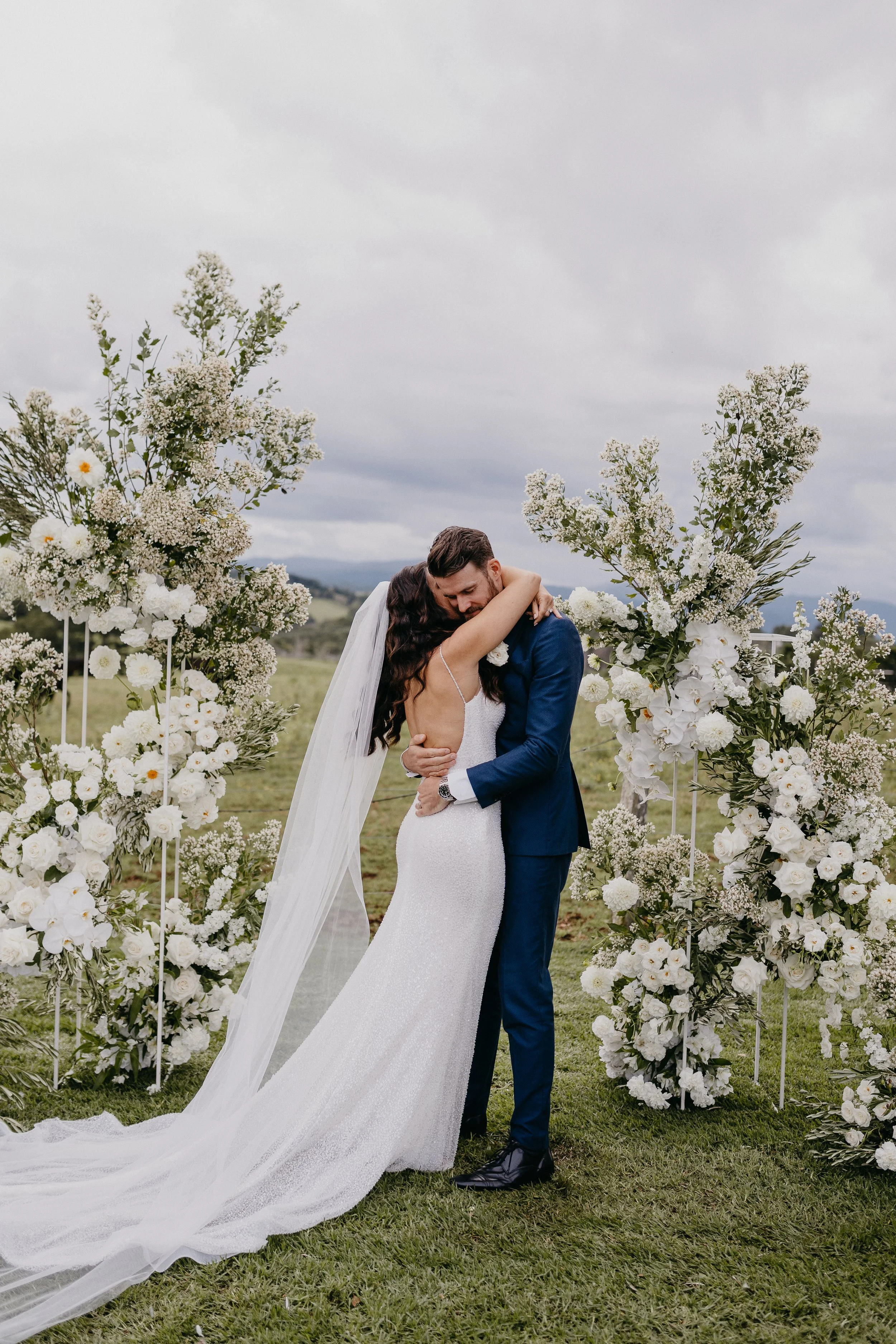 A bride and groom embrace in front of floral wedding decorations outdoors. The bride wears a white gown and veil, and the groom is in a blue suit. Overcast sky in the background.