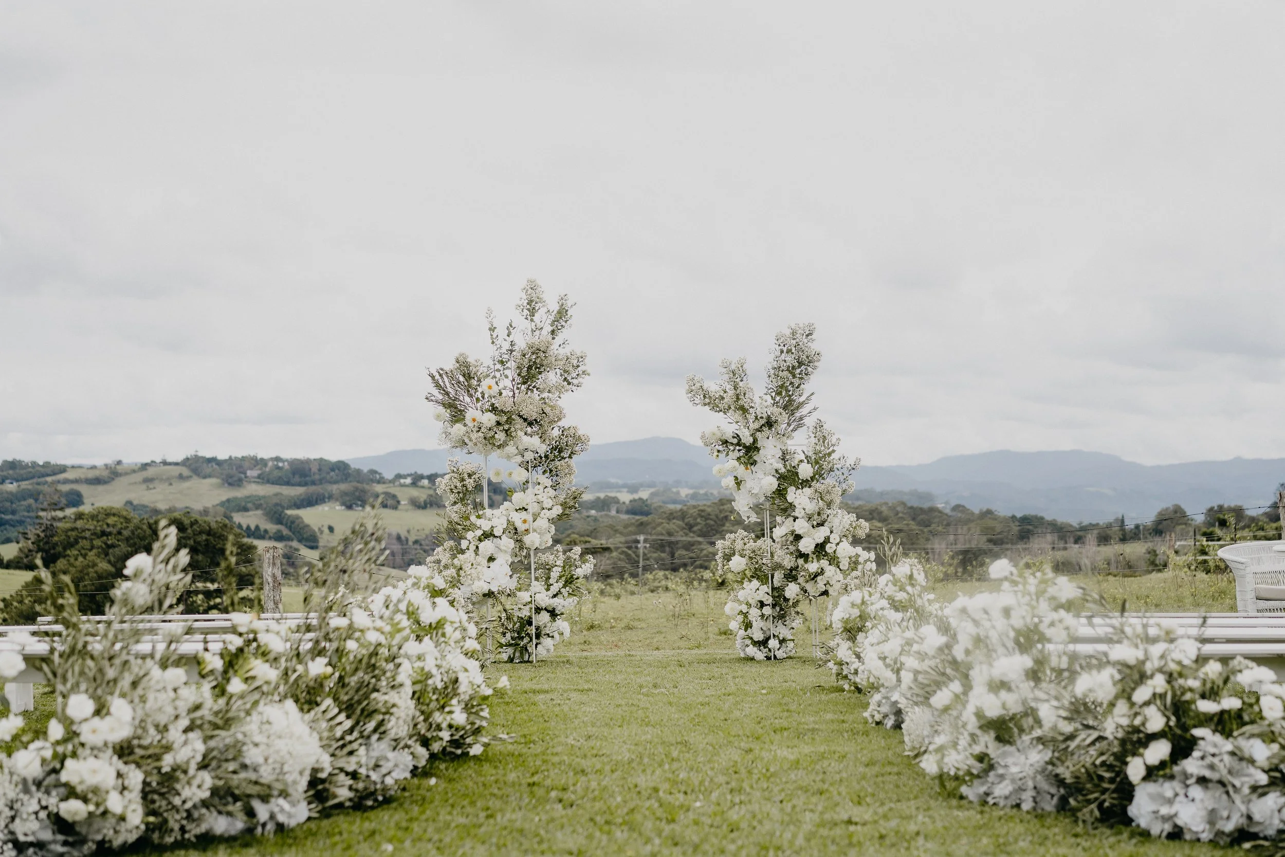 Outdoor wedding altar with white floral arrangements on either side and in the center, set in a green field with mountains in the background under a cloudy sky.