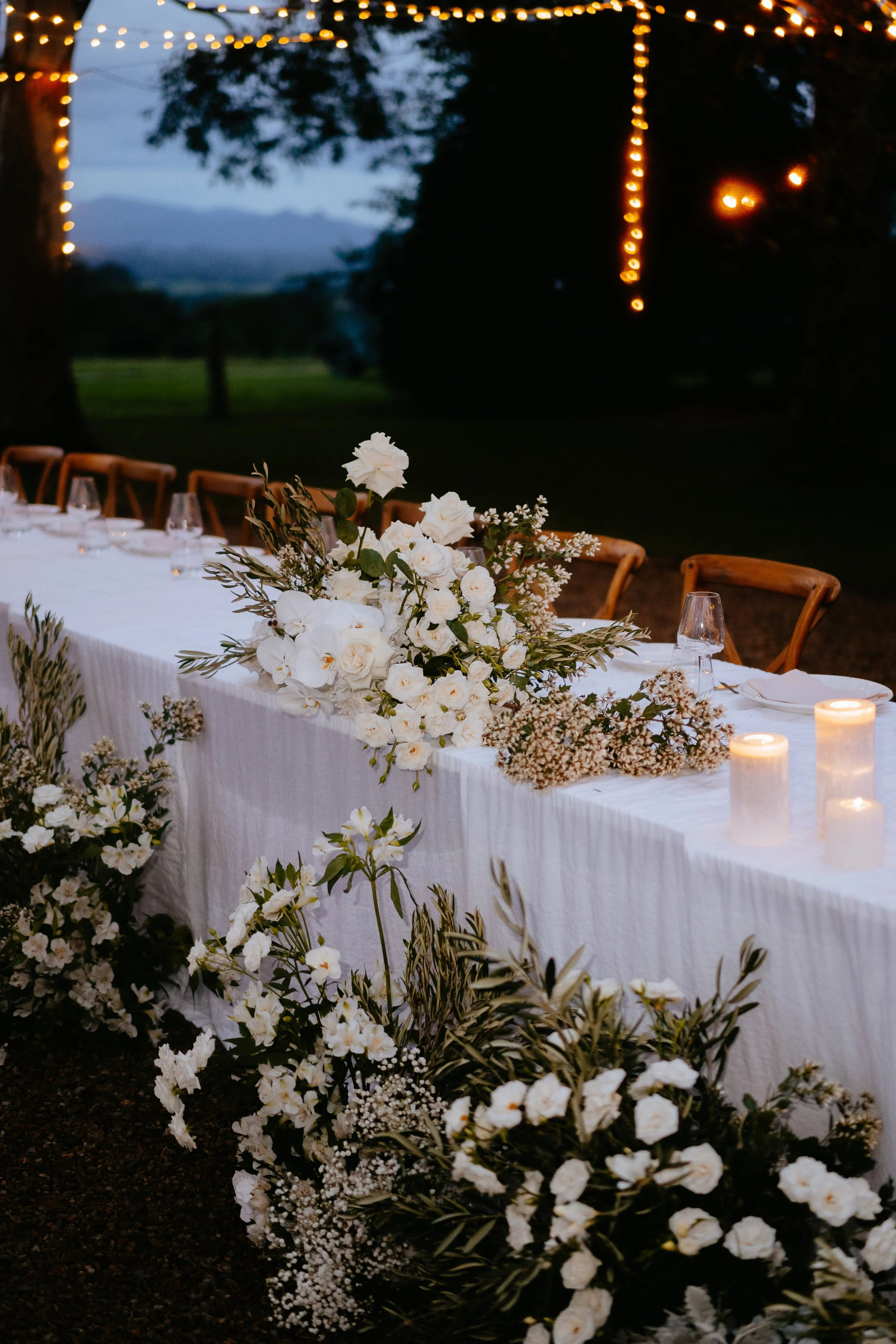 Elegant outdoor wedding reception table decorated with white flowers, greenery, candles, and glassware, set against a backdrop of trees and string lights at dusk.
