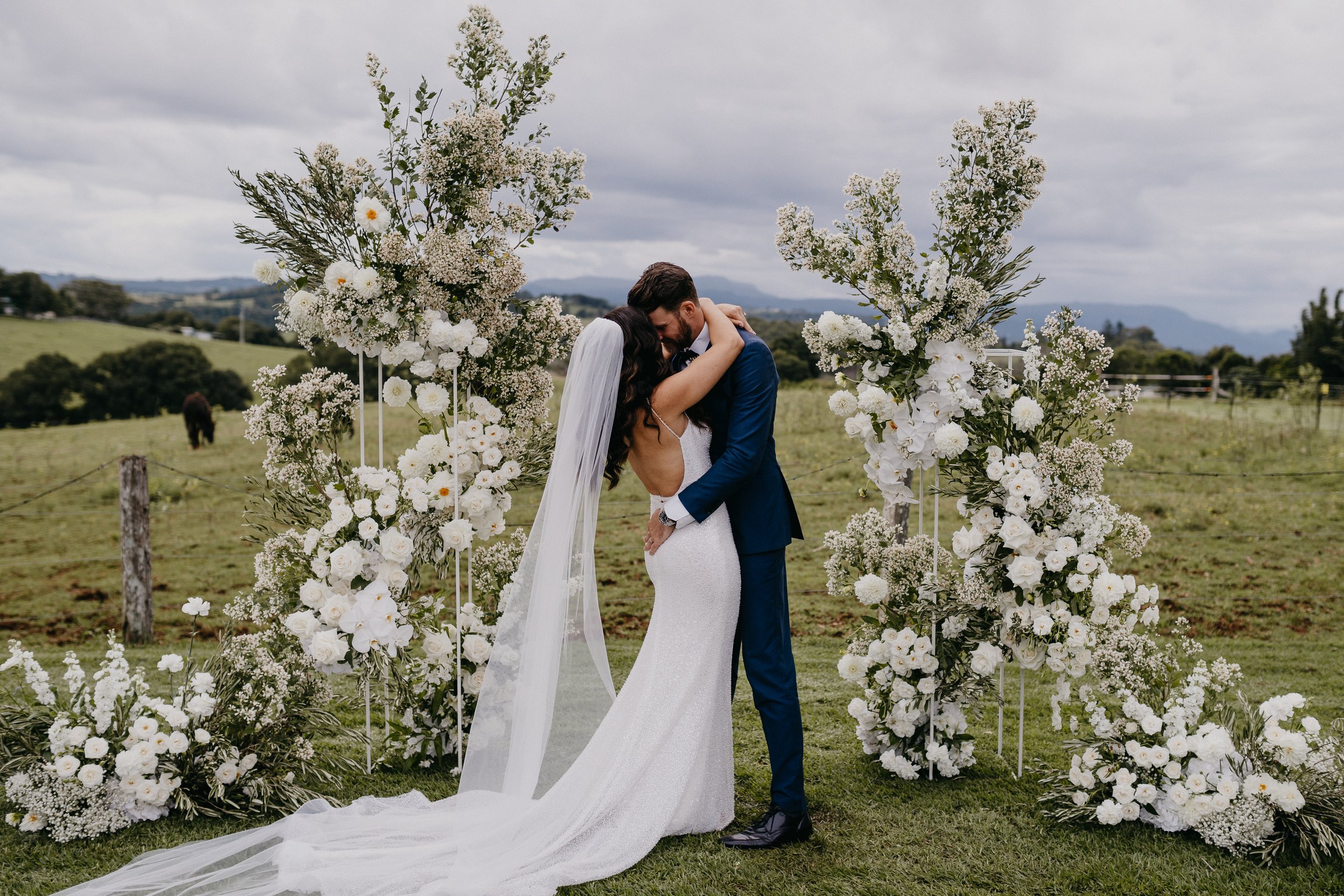 A bride and groom sharing a kiss during their outdoor wedding ceremony surrounded by a floral arch made of white flowers and greenery, with a rural landscape and cloudy sky in the background.