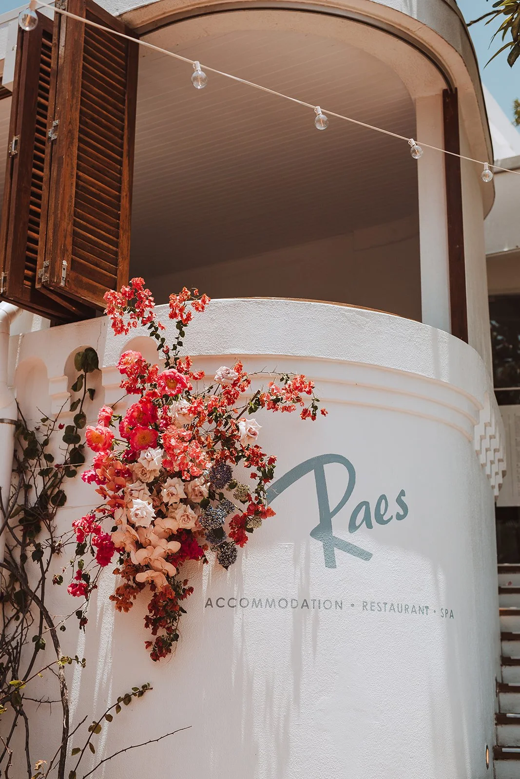 Exterior of a building with a white rounded wall decorated with pink and red flowers and the sign 'Paes' indicating accommodation, restaurant, and spa. There is a string of outdoor lights hanging above.