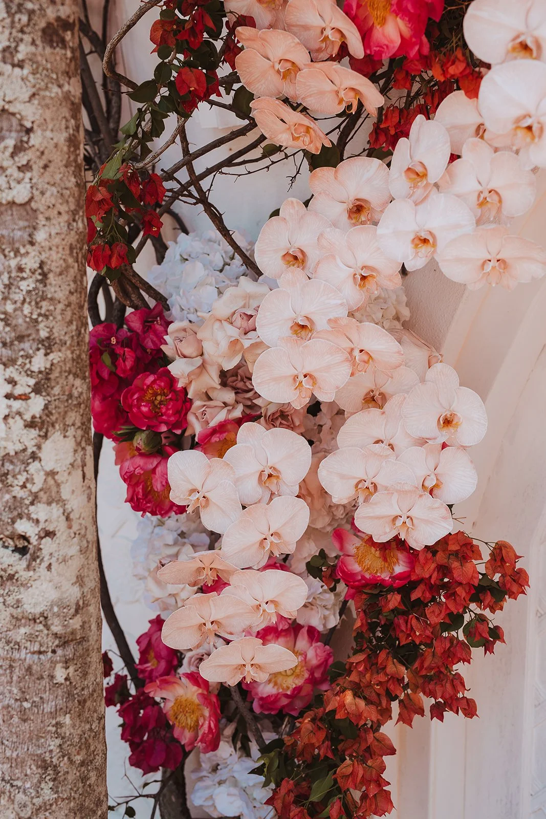 A floral arrangement featuring peach, white, pink, and red flowers along with green leaves and branches, set against a neutral background.